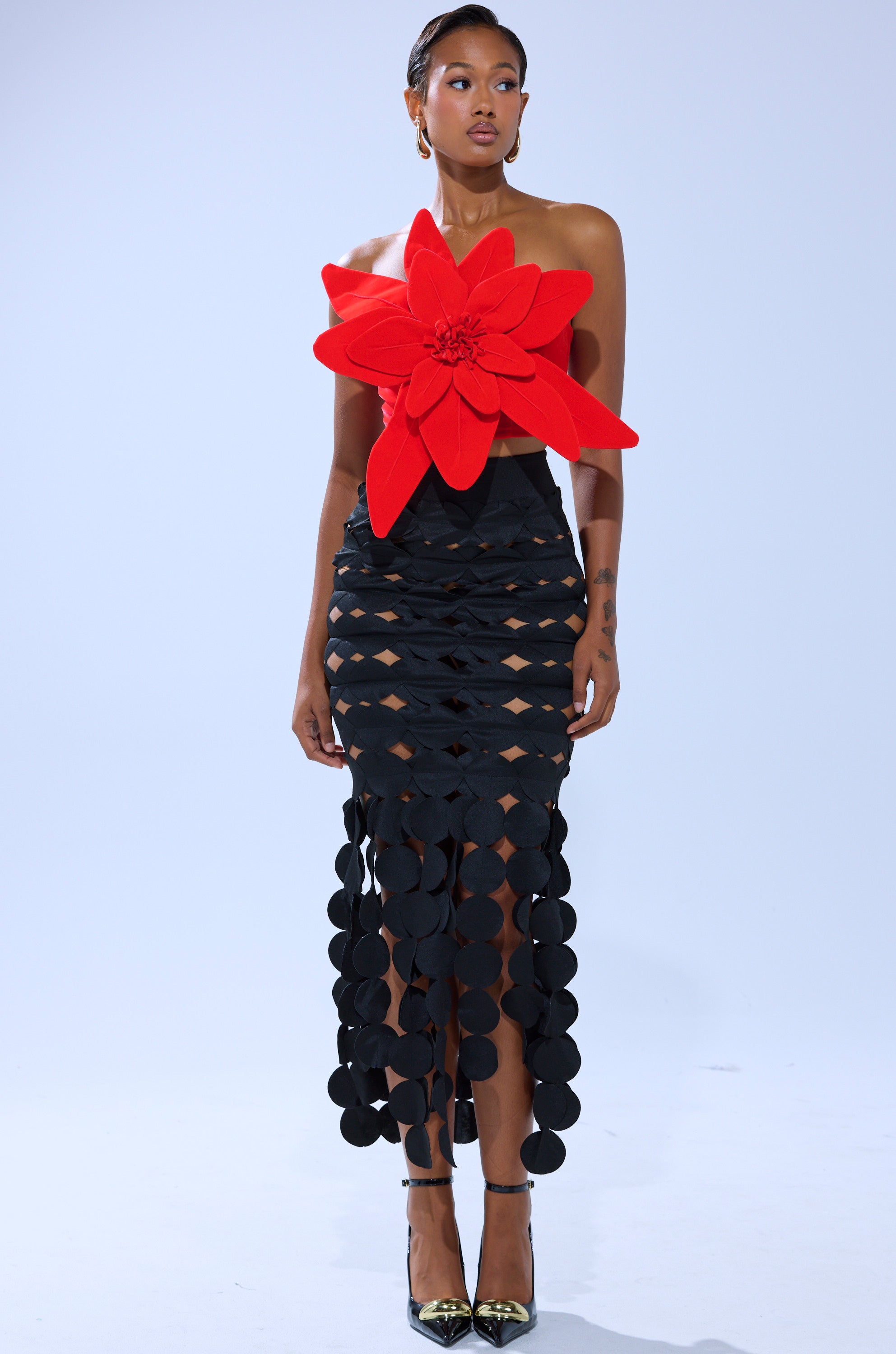 A woman models the HELLA MELODIES FLOWER TOP, a bold red velvet piece with an oversized flower applique, styled with a black circular-cutout skirt and metallic pointed-toe heels against a plain light backdrop.