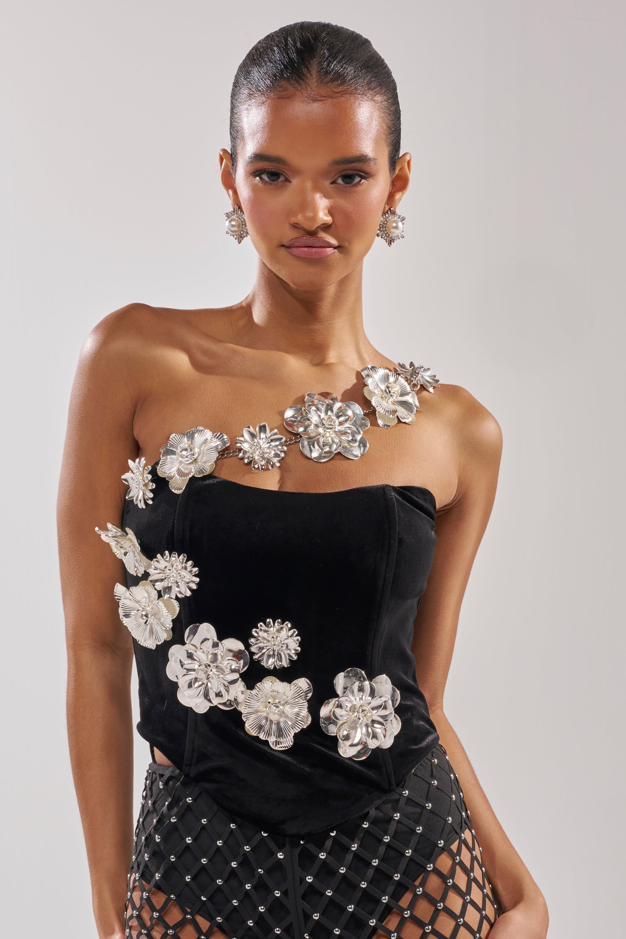 A woman wears the AKIRA BLINGING OUT CORSET, a strapless black top with metallic floral embellishments and chain link details. She pairs it with statement floral earrings and a black mesh skirt, her hair pulled back against a plain background. 0
