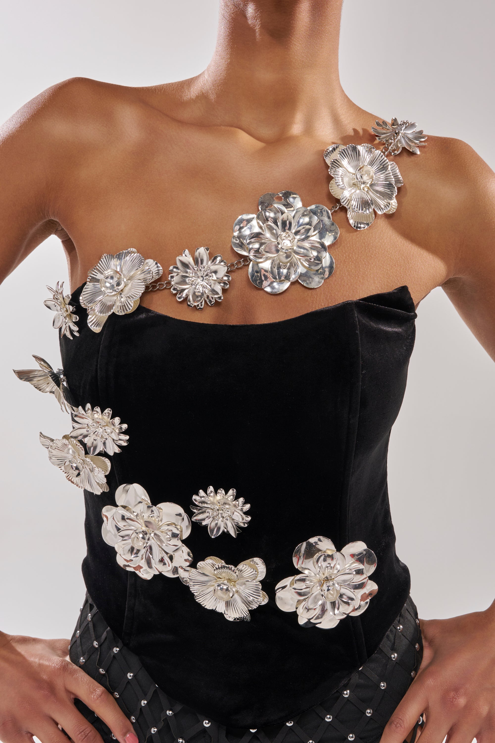 A person wearing the BLINGING OUT CORSET—a black velvet strapless top with bold, shiny silver floral embellishments—stands with hands on hips against a plain background. 11