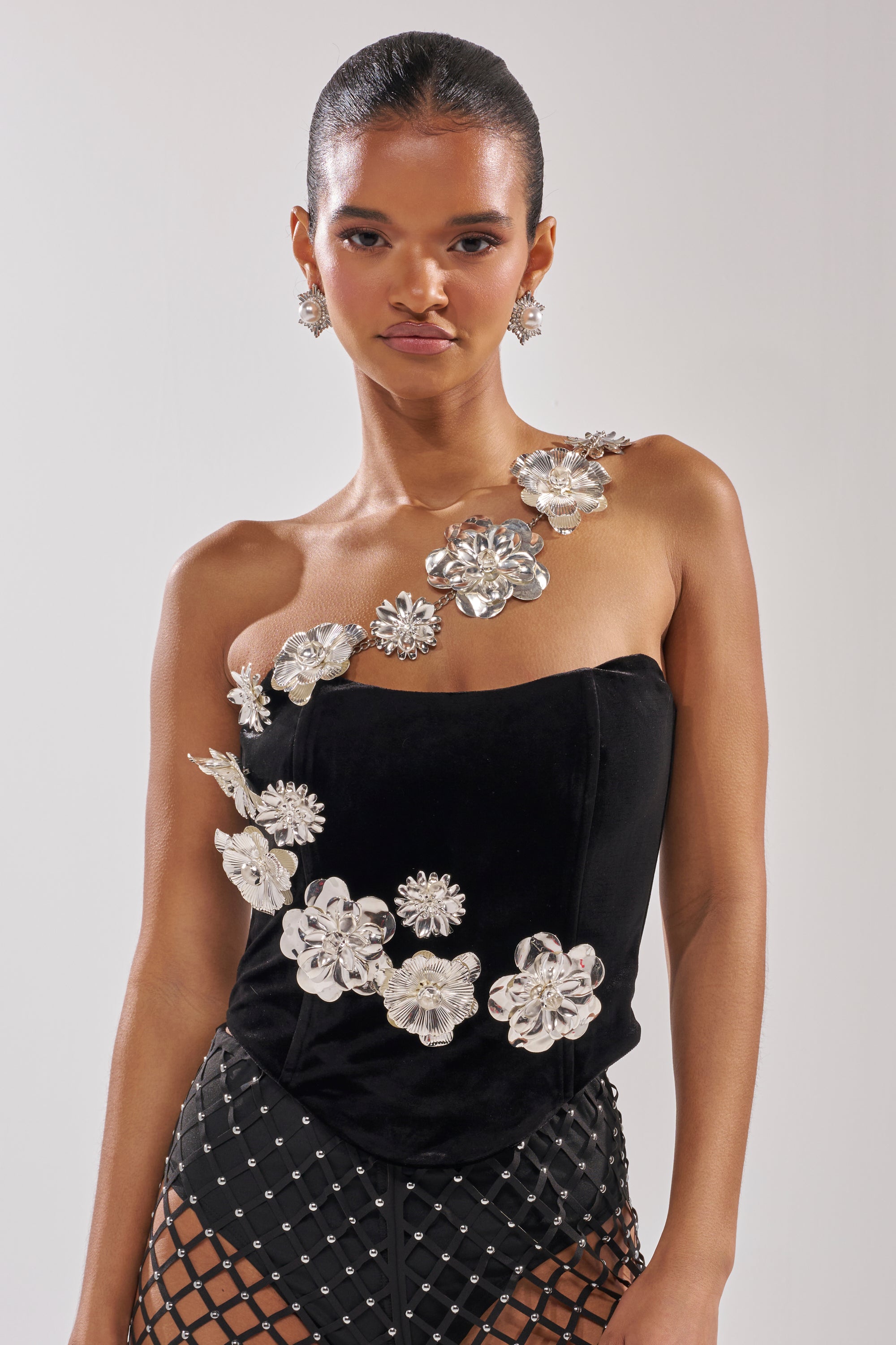 A woman wears the BLINGING OUT CORSET in black stretch velvet with silver floral embellishments, paired with a black skirt featuring a diamond-pattern mesh overlay, standing against a plain light background. 1