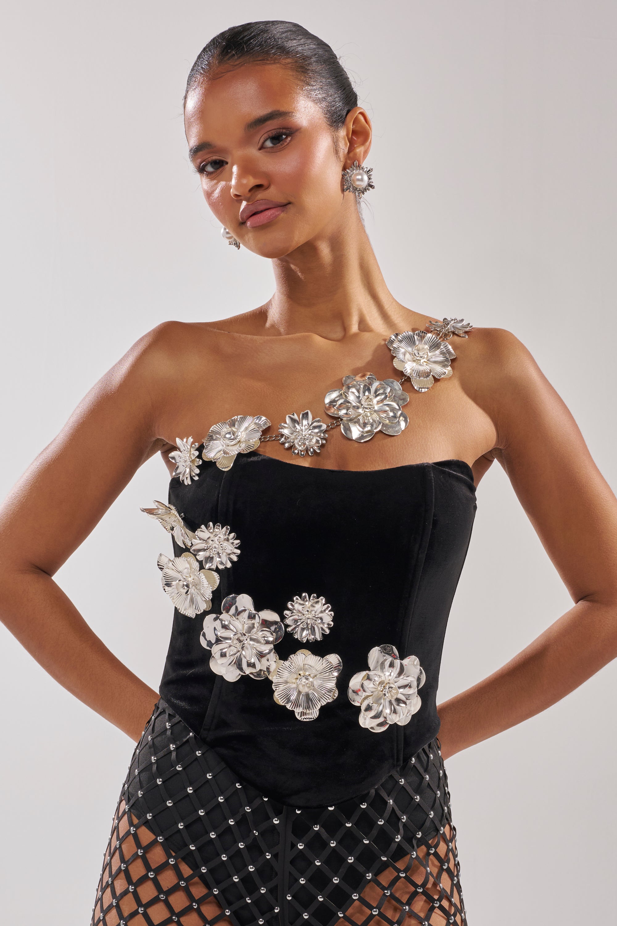 A woman models the BLINGING OUT CORSET—a strapless stretch velvet top with bold silver flower appliqués—paired with a black mesh skirt, slicked-back hair, and matching silver flower earrings. 2