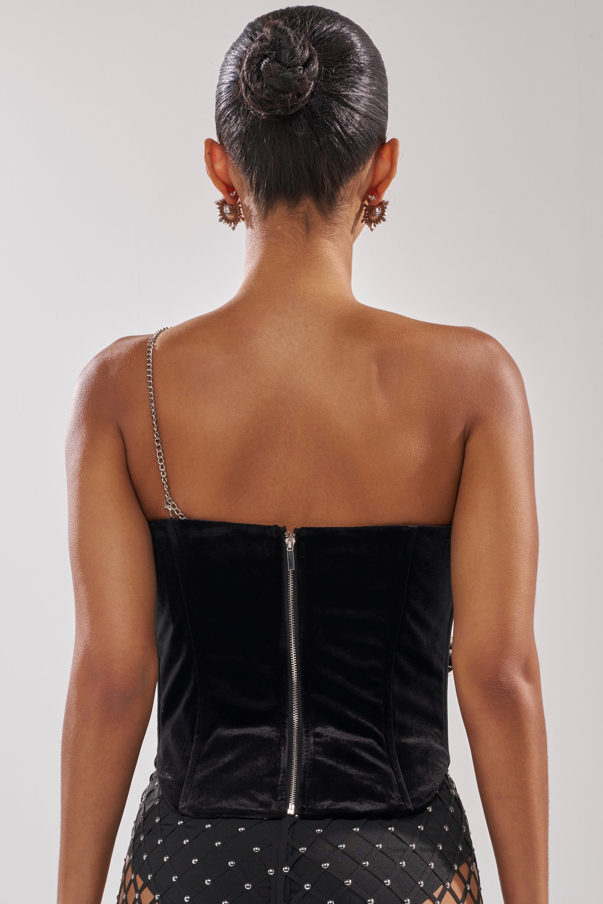 A woman is seen from behind wearing the BLINGING OUT CORSET—a black stretch velvet, one-shoulder top with chain link accents and a silver back zipper—her hair in a neat bun with earrings, styled with patterned pants. 5