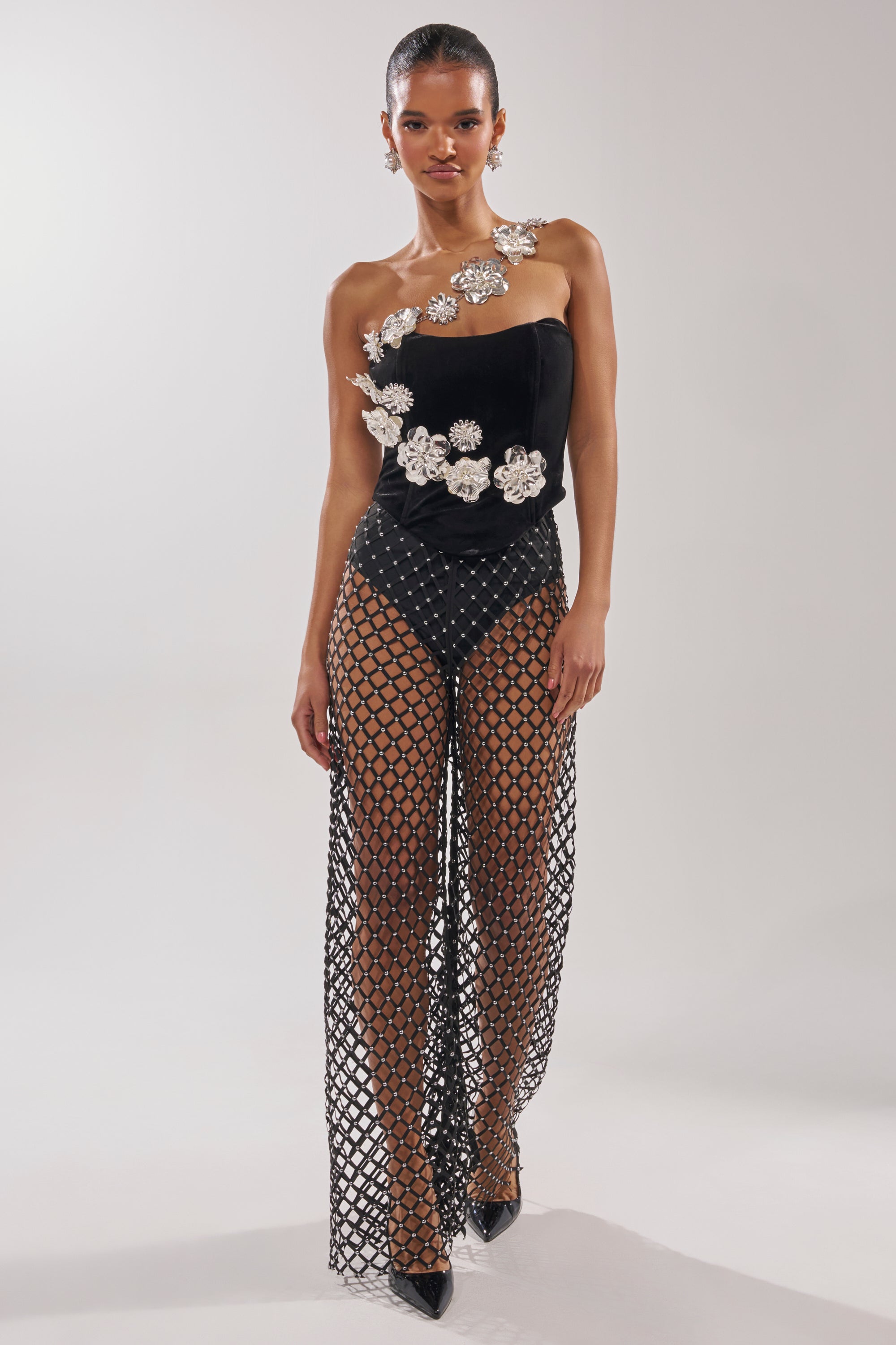 A woman models the BLINGING OUT CORSET, a unique black strapless piece with silver floral appliqués, paired with wide-leg geometric netted pants over high-waisted black briefs. 7