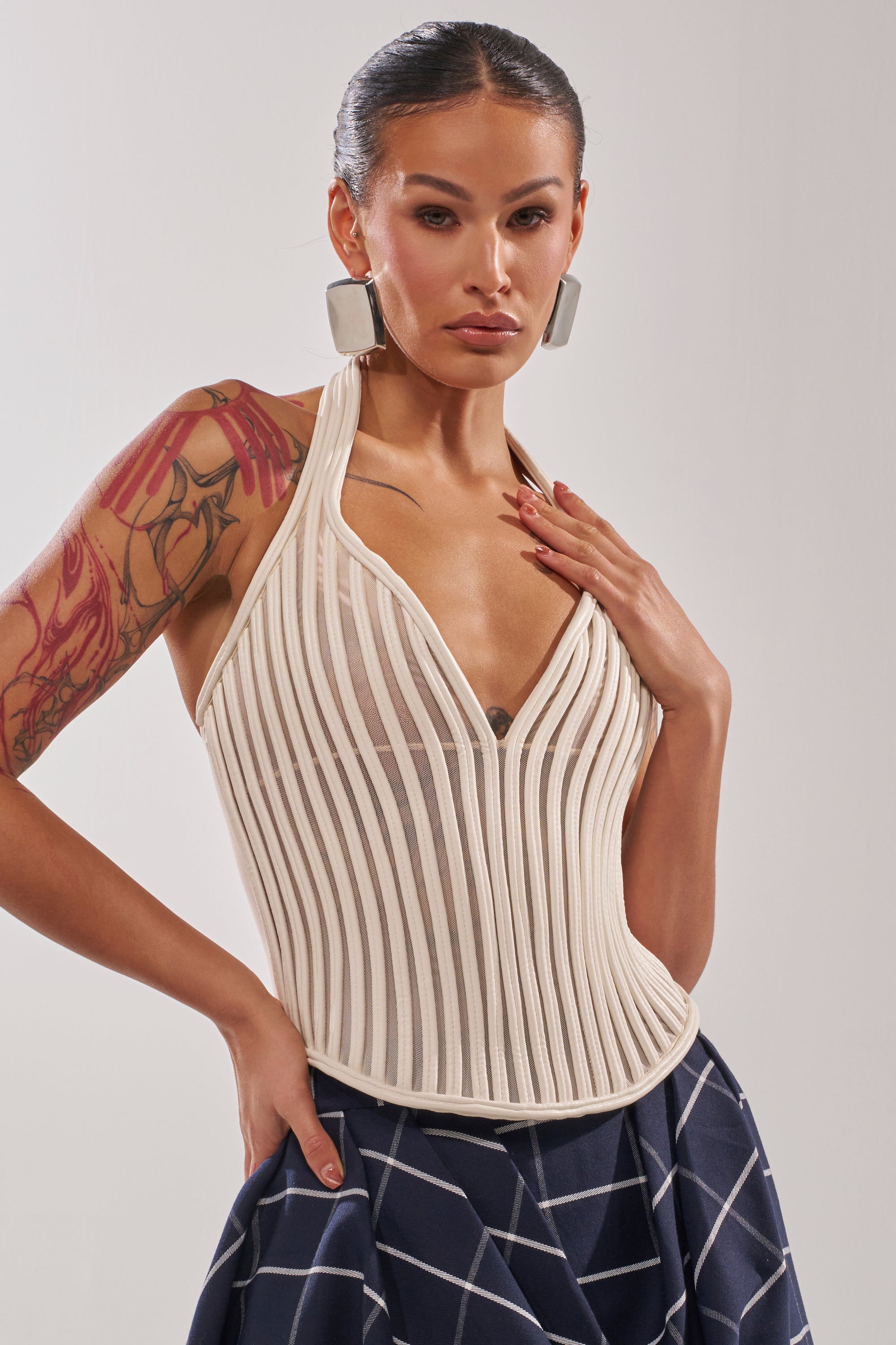 A confident woman with slicked-back hair wears the GET GROOVY WITH IT MESH CORSET, a halter-neck sheer top, paired with large square earrings and a blue skirt with a white grid. She shows off a dragon tattoo on her left arm against a plain backdrop. 1