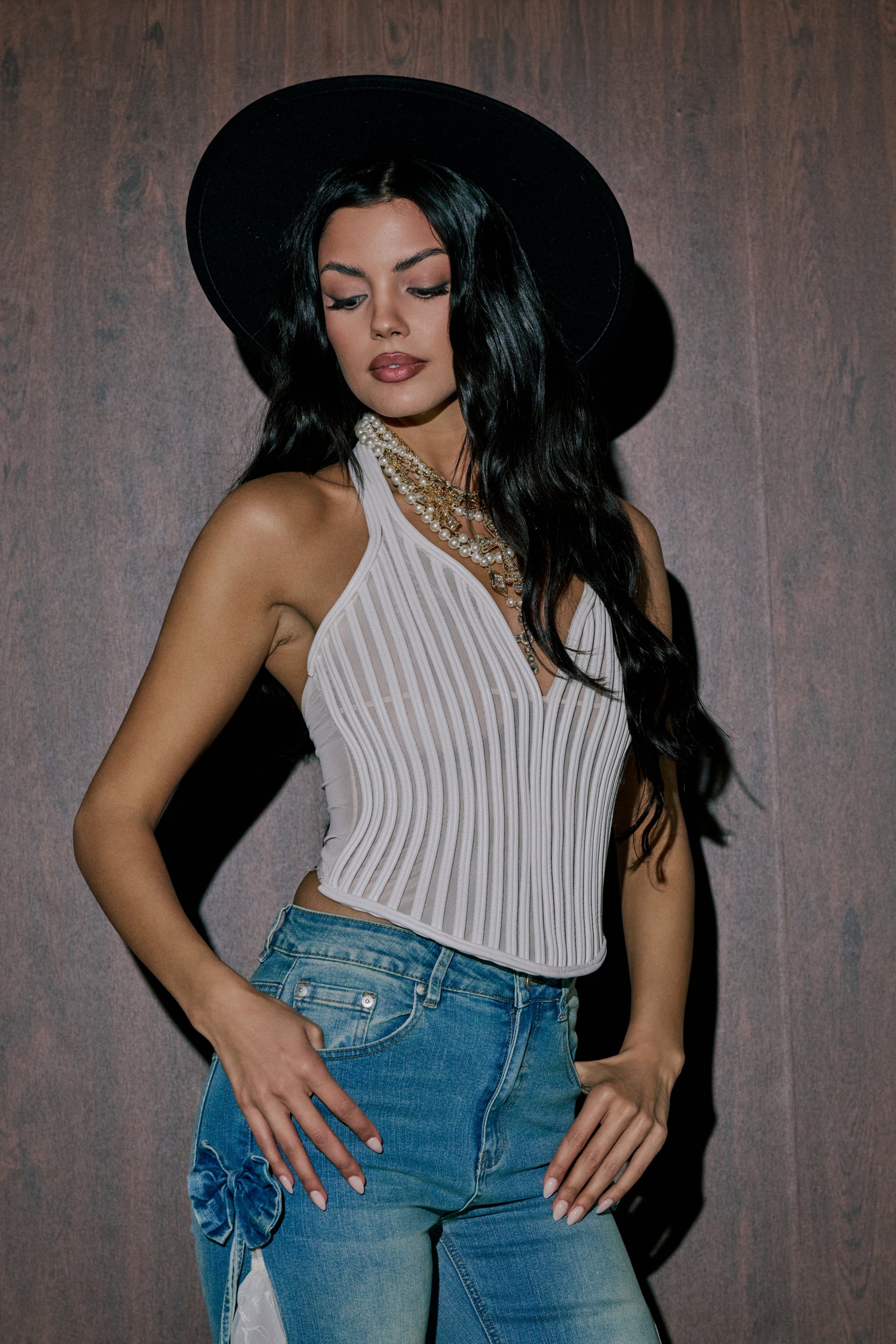 Wearing the GET GROOVY WITH IT MESH CORSET, a woman with long dark hair and a black wide-brim hat pairs layered necklaces and blue jeans, standing against a dark wooden background while looking down to her left. 0