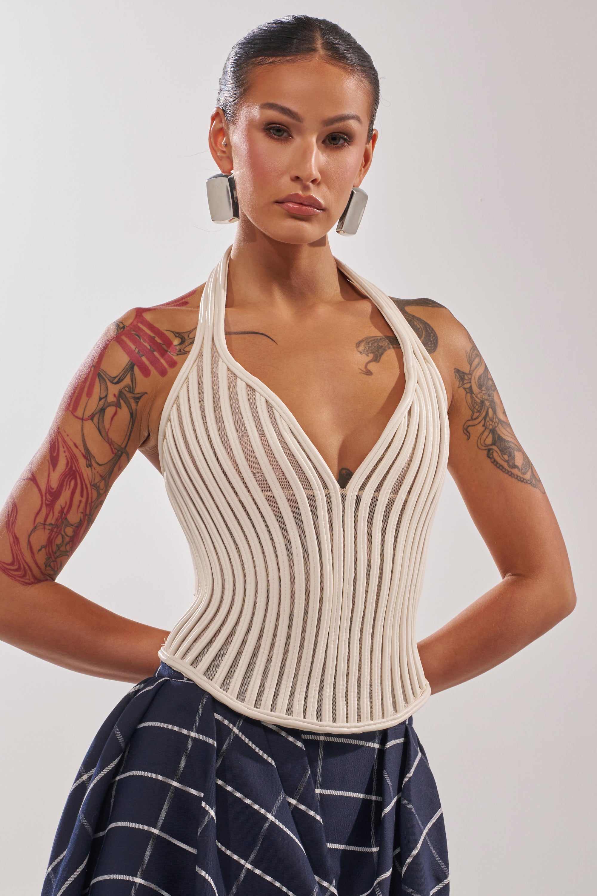 A woman with dark hair in a sleek bun wears large square earrings and the GET GROOVY WITH IT MESH CORSET, a cream structured halter top with sculpted fit and vertical lines, paired with a navy grid-patterned skirt. Arm tattoos show her confidence. 3