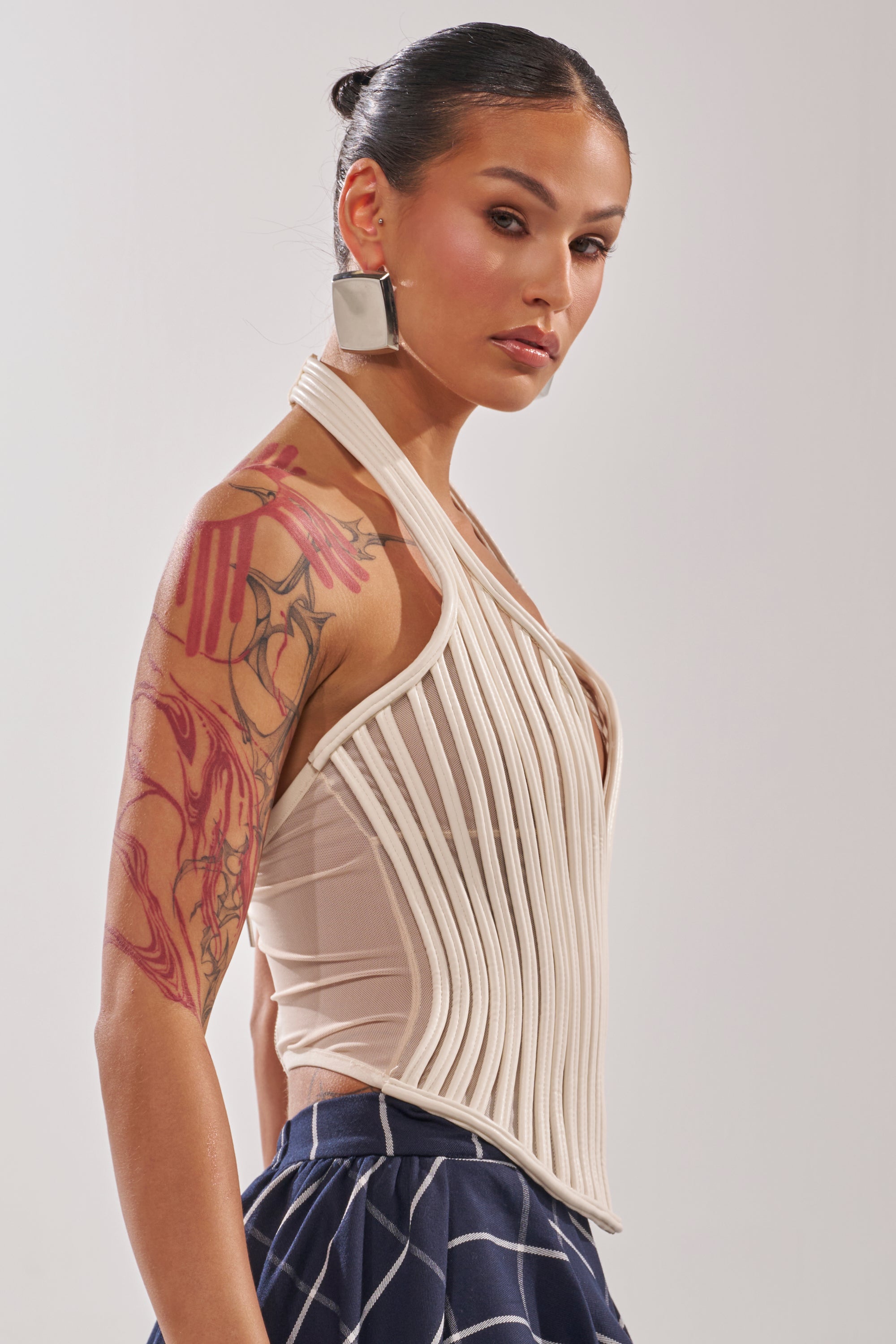 A person with slicked-back hair, bold earrings, and a red arm tattoo wears the GET GROOVY WITH IT MESH CORSET featuring a cream halter neckline and sculpted fit, paired with navy pants with white lines against a plain background. 4