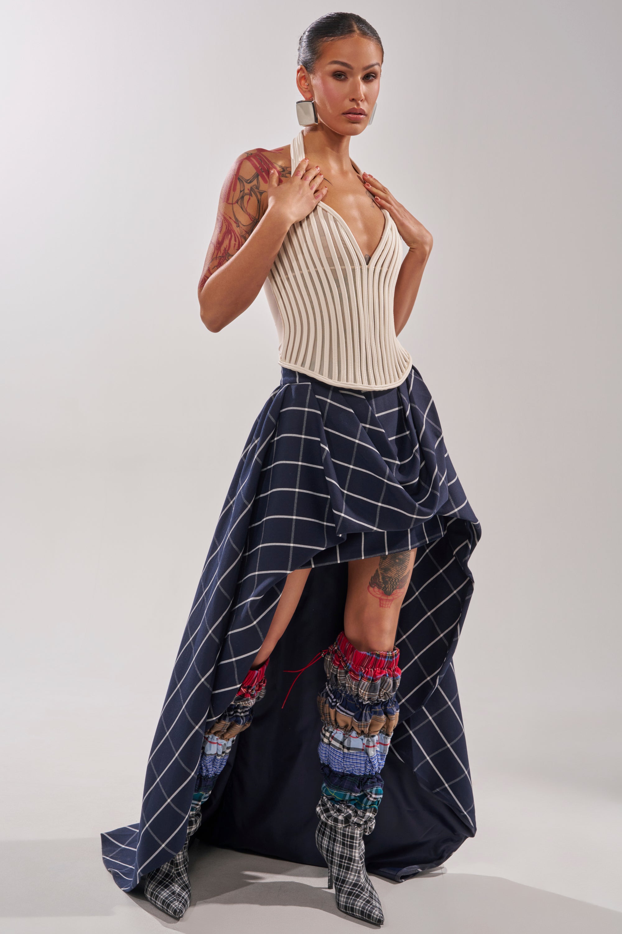 A woman poses in the GET GROOVY WITH IT MESH CORSET, a cream-colored halter-style top, paired with an asymmetrical navy skirt featuring a white grid pattern, colorful textured leg warmers, and plaid boots against a plain light background. 7