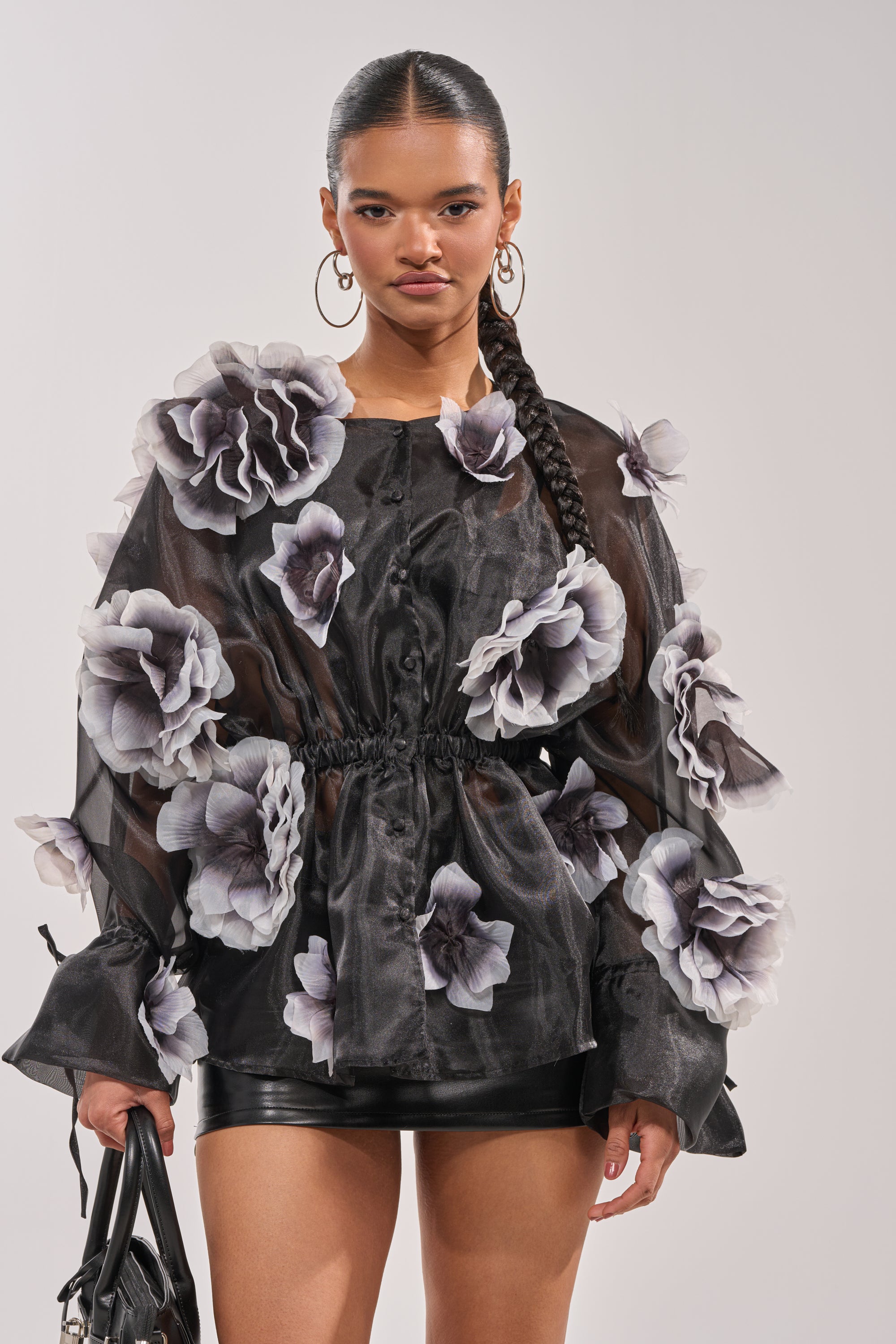 A woman with a long braided ponytail wears the DARK BLOOM CHIFFON BLOUSE, featuring large white and gray 3D floral appliqués. She pairs it with black shorts, hoop earrings, and carries a sleek black handbag. 1