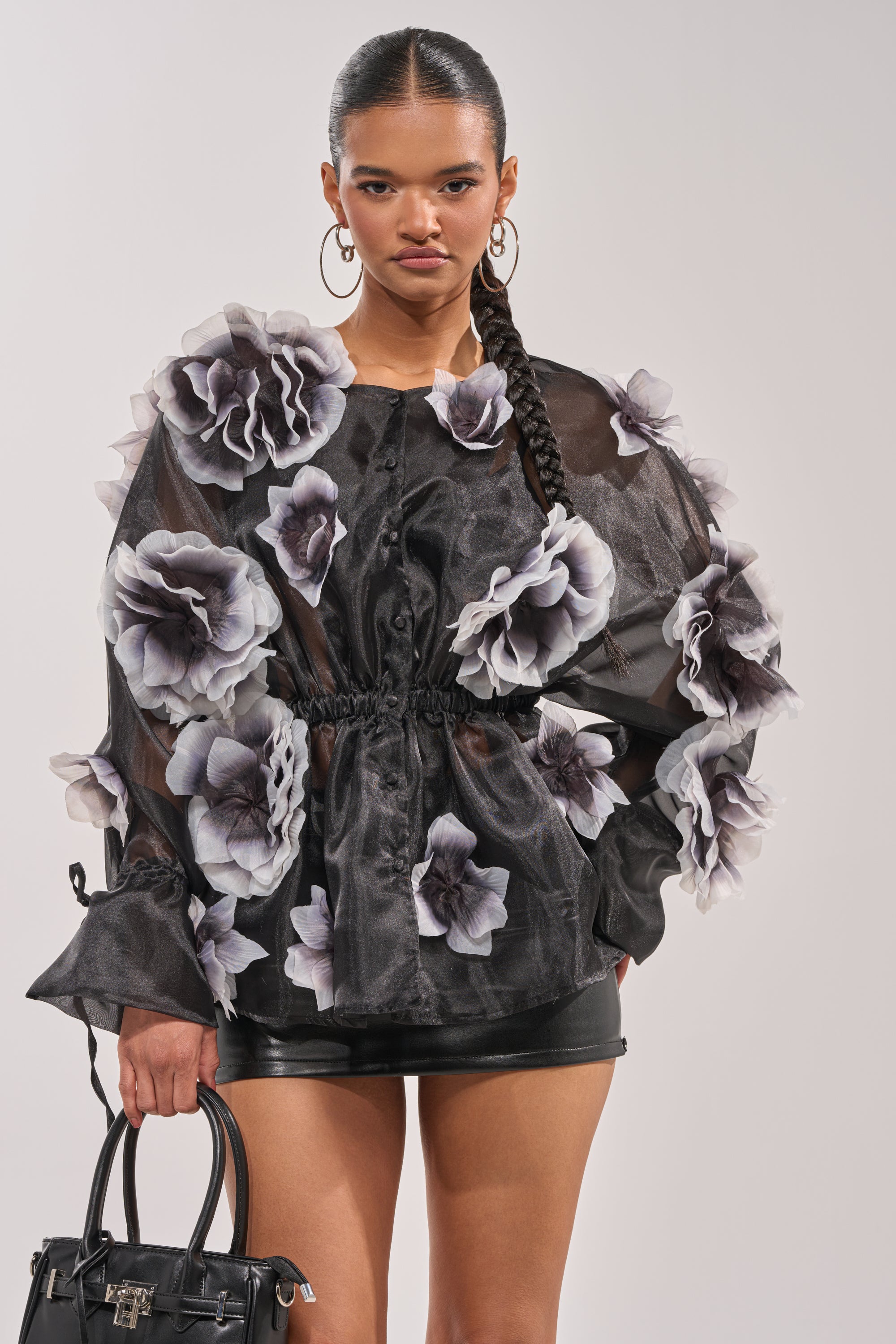 A woman in the DARK BLOOM CHIFFON BLOUSE with 3D floral appliqués, a black leather mini skirt, hoop earrings, and a black handbag stands against a plain background. 2