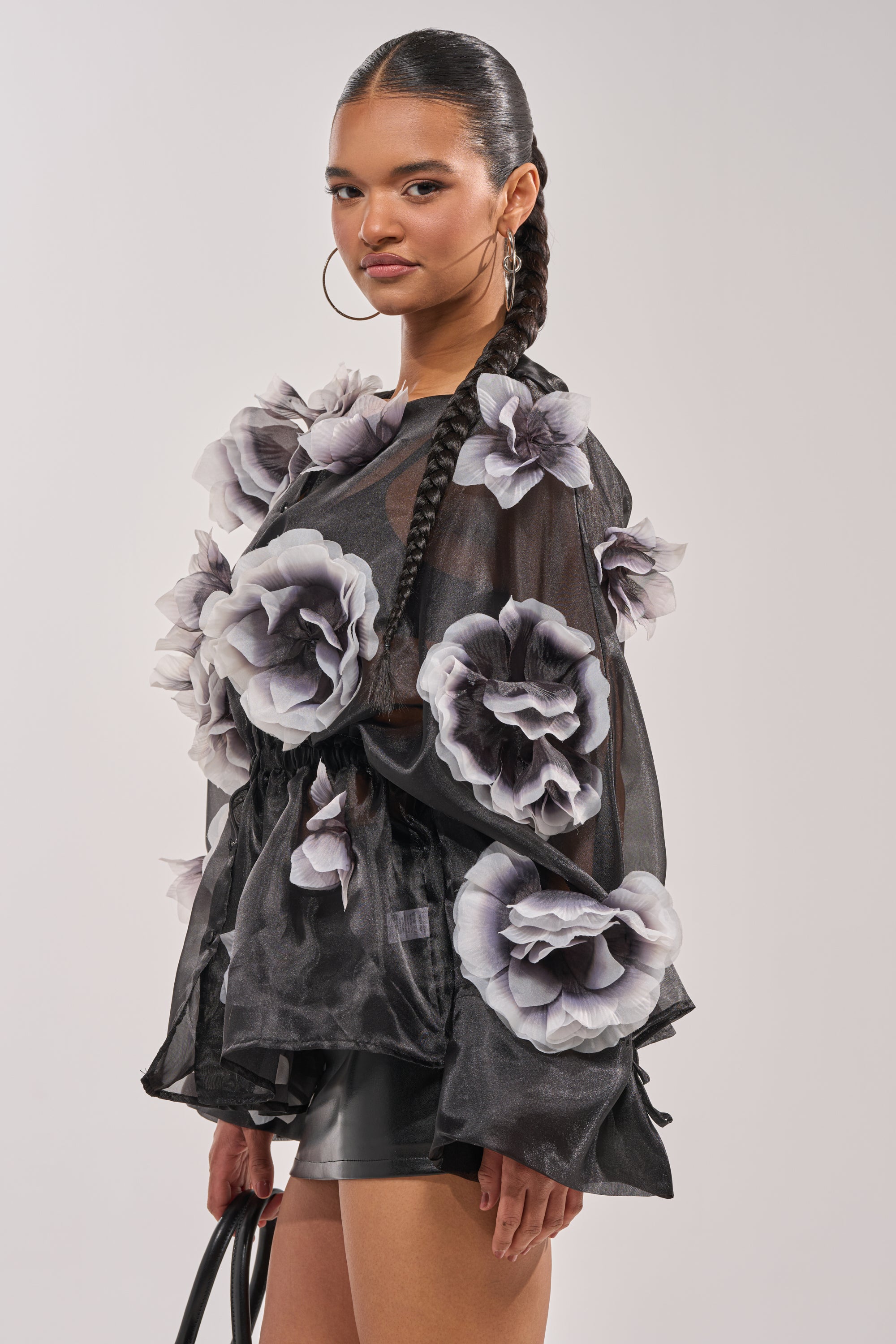A woman with a long braid wears the DARK BLOOM CHIFFON BLOUSE, a sheer black top adorned with large fabric flowers, paired with a black mini skirt and hoop earrings. She holds a black bag and poses confidently against a plain backdrop. 3