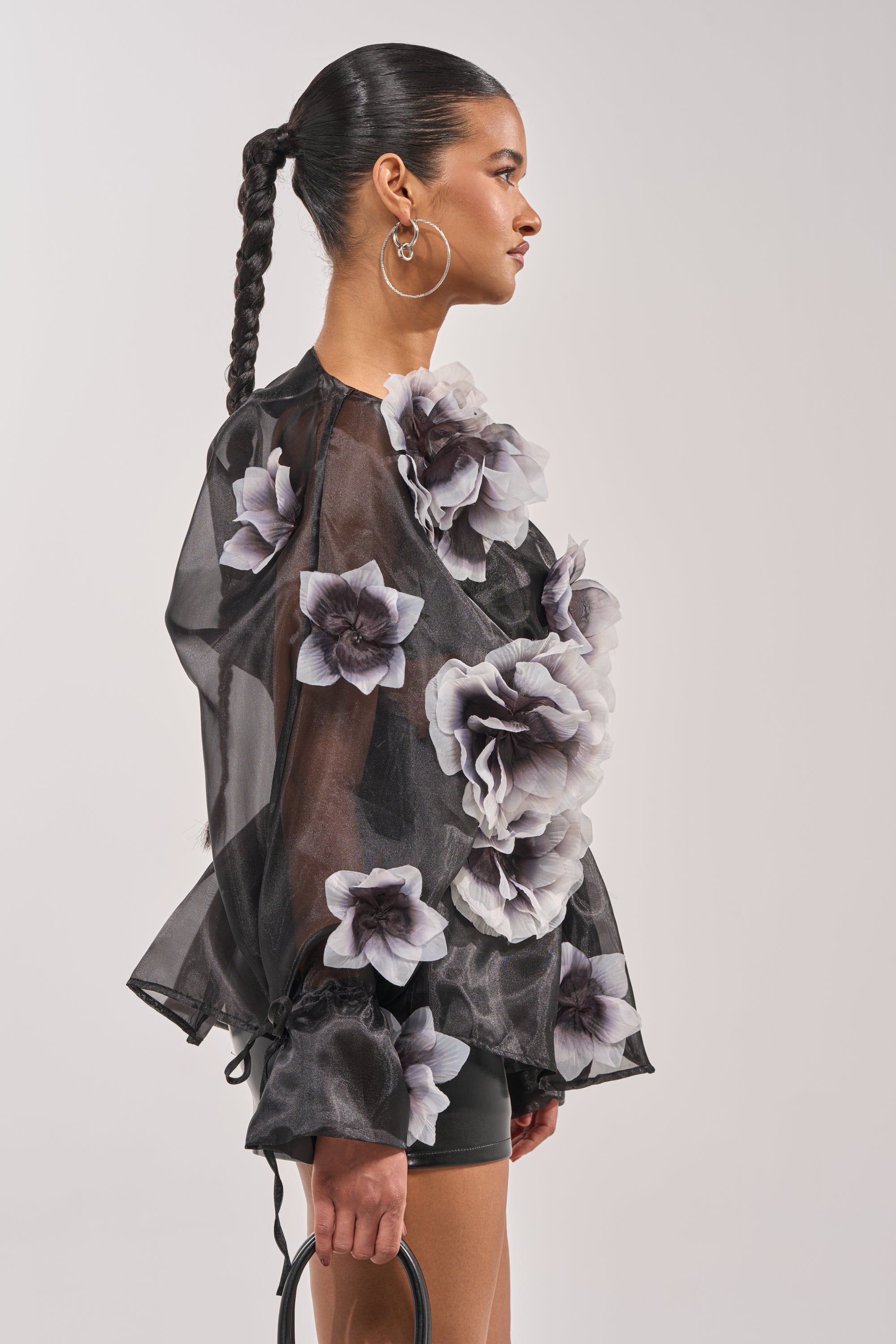 A woman with a slick, braided ponytail wears large hoop earrings and the DARK BLOOM CHIFFON BLOUSE featuring sheer black fabric and bold 3D floral appliqués. She holds a black handbag, standing against a plain background. 4