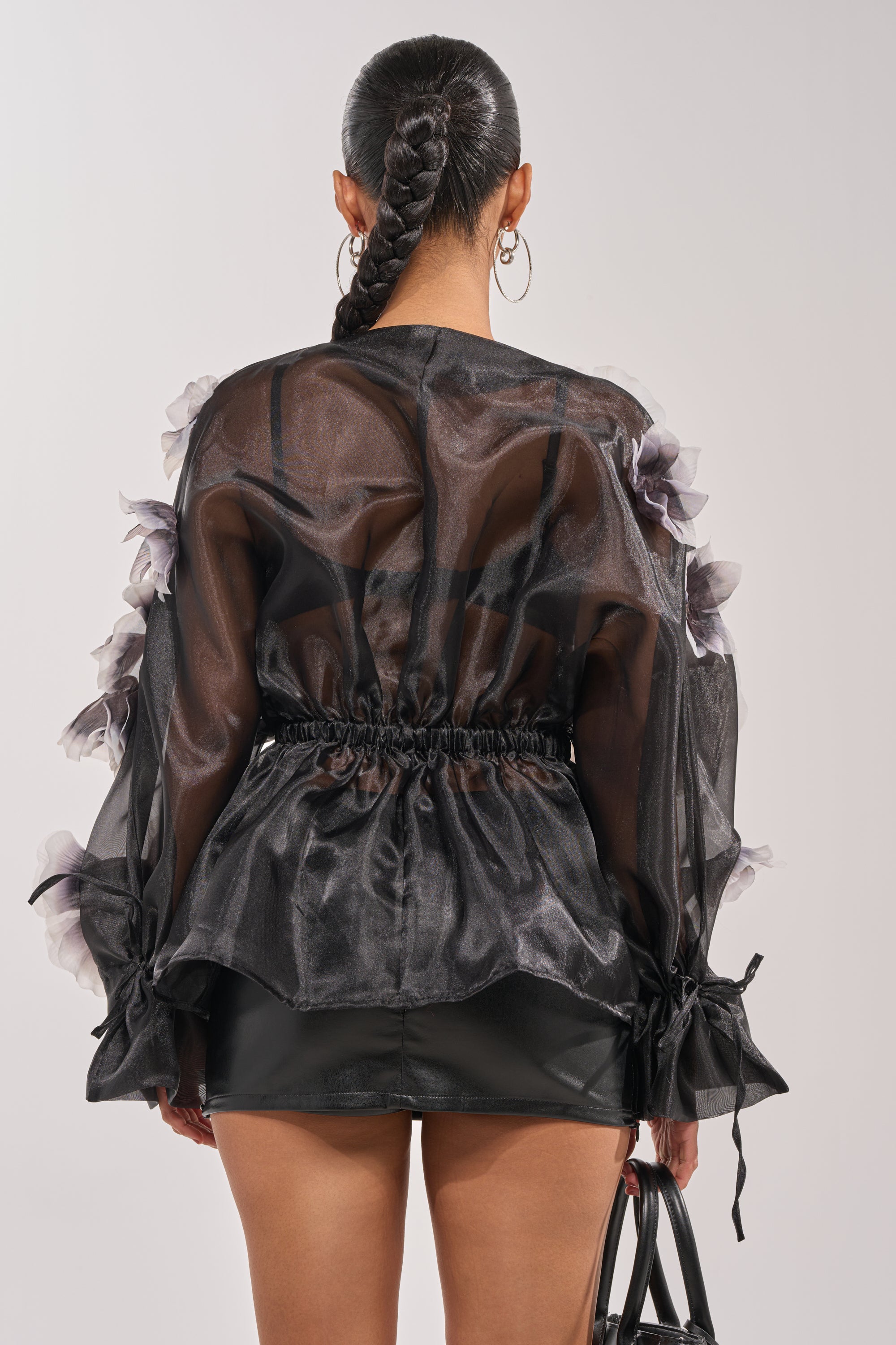 A woman with a long braid is seen from the back, wearing the DARK BLOOM CHIFFON BLOUSE—a sheer black top with floral details and a cinched waist—paired with a black leather mini skirt and holding a black handbag. 5
