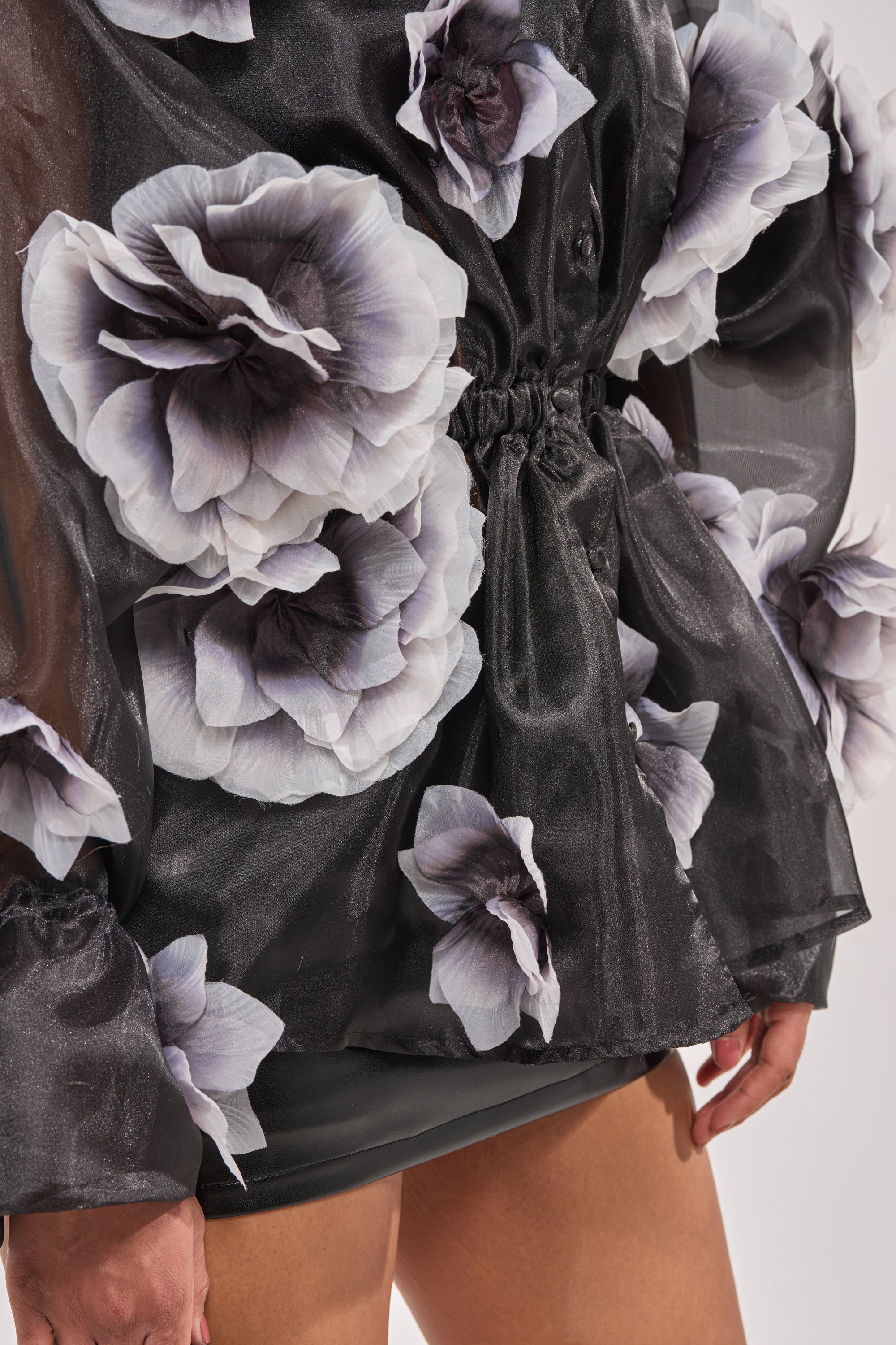 A close-up of a person wearing the DARK BLOOM CHIFFON BLOUSE, featuring sheer black fabric with large grayish-white 3D floral appliqué and voluminous sleeves, styled elegantly with a dark skirt. 8