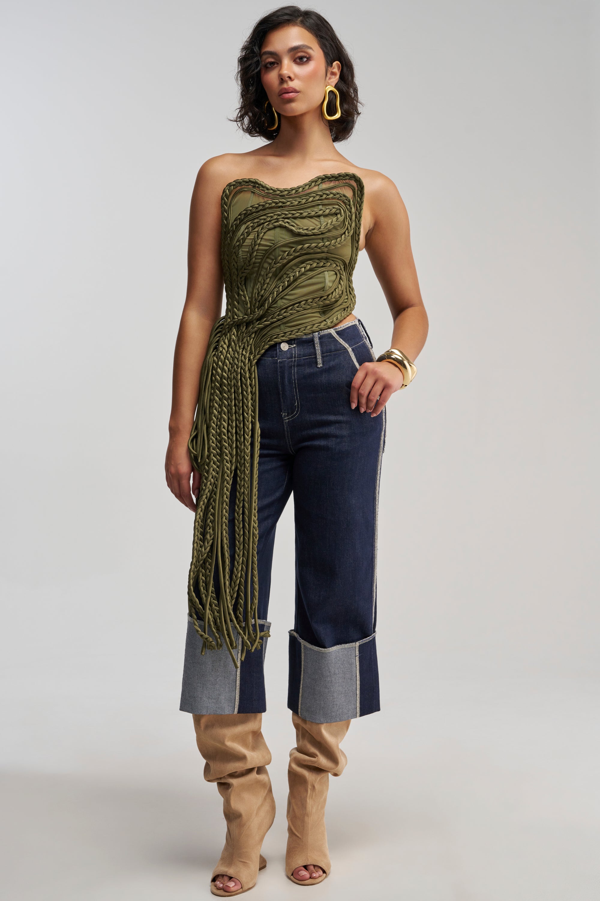 Wearing the TANGLED INTENTIONS CORSET—a green, textured top with long fringe—styled as a festival outfit, a woman poses against a plain background. She pairs the corset with dark cuffed jeans, tan slouchy boots, and bold gold jewelry for a standout look.