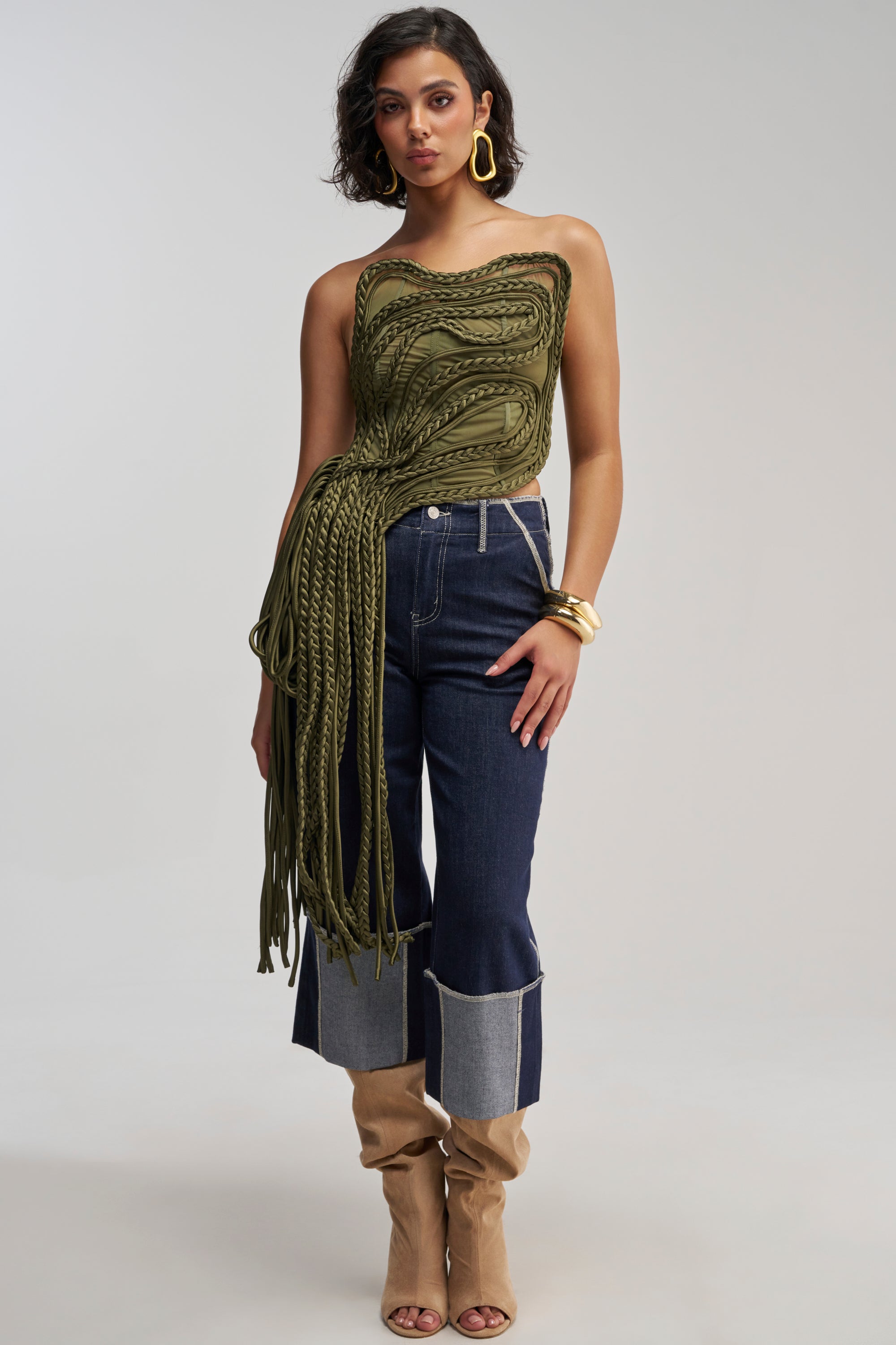 A woman poses in the green, textured TANGLED INTENTIONS CORSET with an asymmetrical rope design and long fringe, paired with dark denim jeans featuring light cuffs, tan heeled boots, gold hoop earrings, and bracelets against a plain background for a fresh streetwear look.