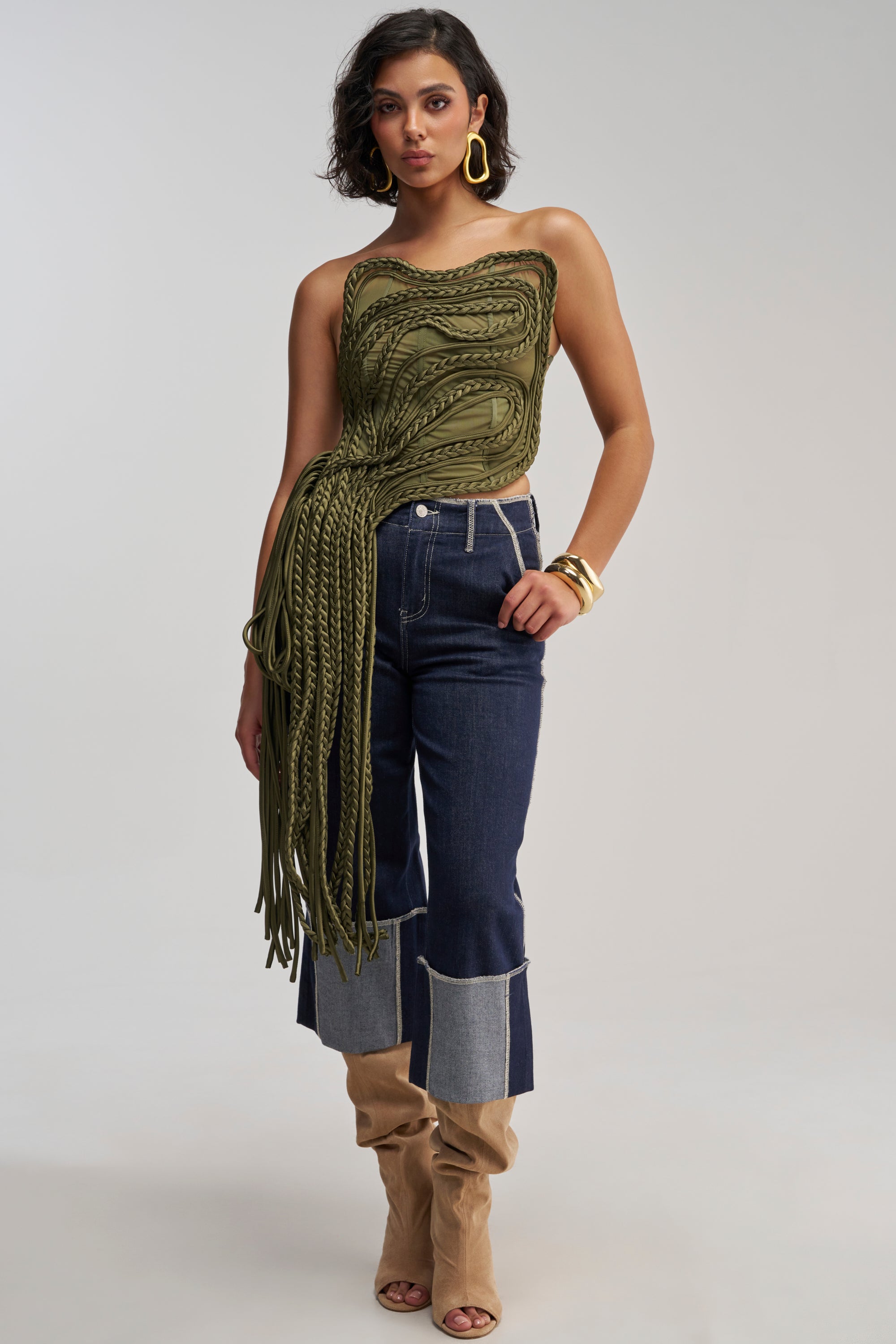 Wearing the TANGLED INTENTIONS CORSET—a striking green, braided, asymmetrical rope corset top with long fringe—paired with dark cropped jeans featuring light cuffs, tan open-toe boots, gold hoop earrings, and bracelets, a woman poses against a plain background for an effortless streetwear look that’s perfect for standing out at a festival.