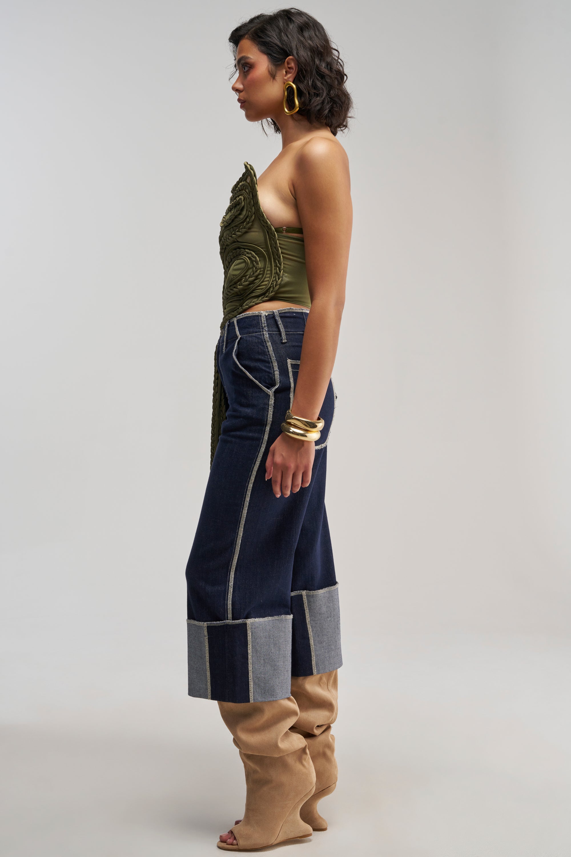 Woman standing sideways in a studio wearing the TANGLED INTENTIONS CORSET—an olive green asymmetrical top—styled with festival outfit energy, blue wide-leg cuffed jeans, tan slouchy boots, gold hoop earrings, and bangles against a plain light background.