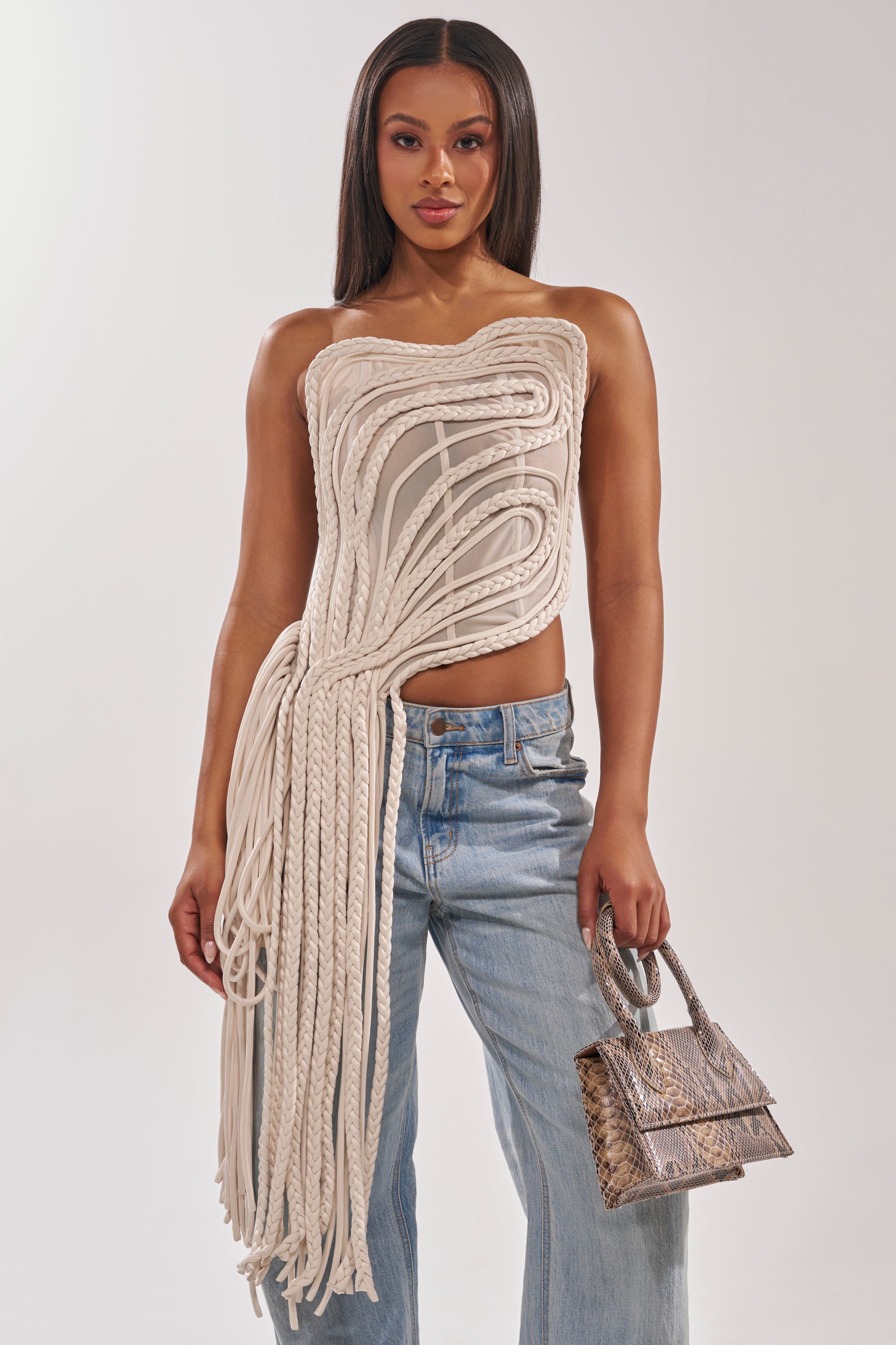 A woman stands against a plain background in the runway-inspired CUTTING YOU OFF MESH ROPE CORSET top, featuring sculpted cream mesh and long asymmetrical rope details, paired with light blue jeans and a small snakeskin-patterned handbag.