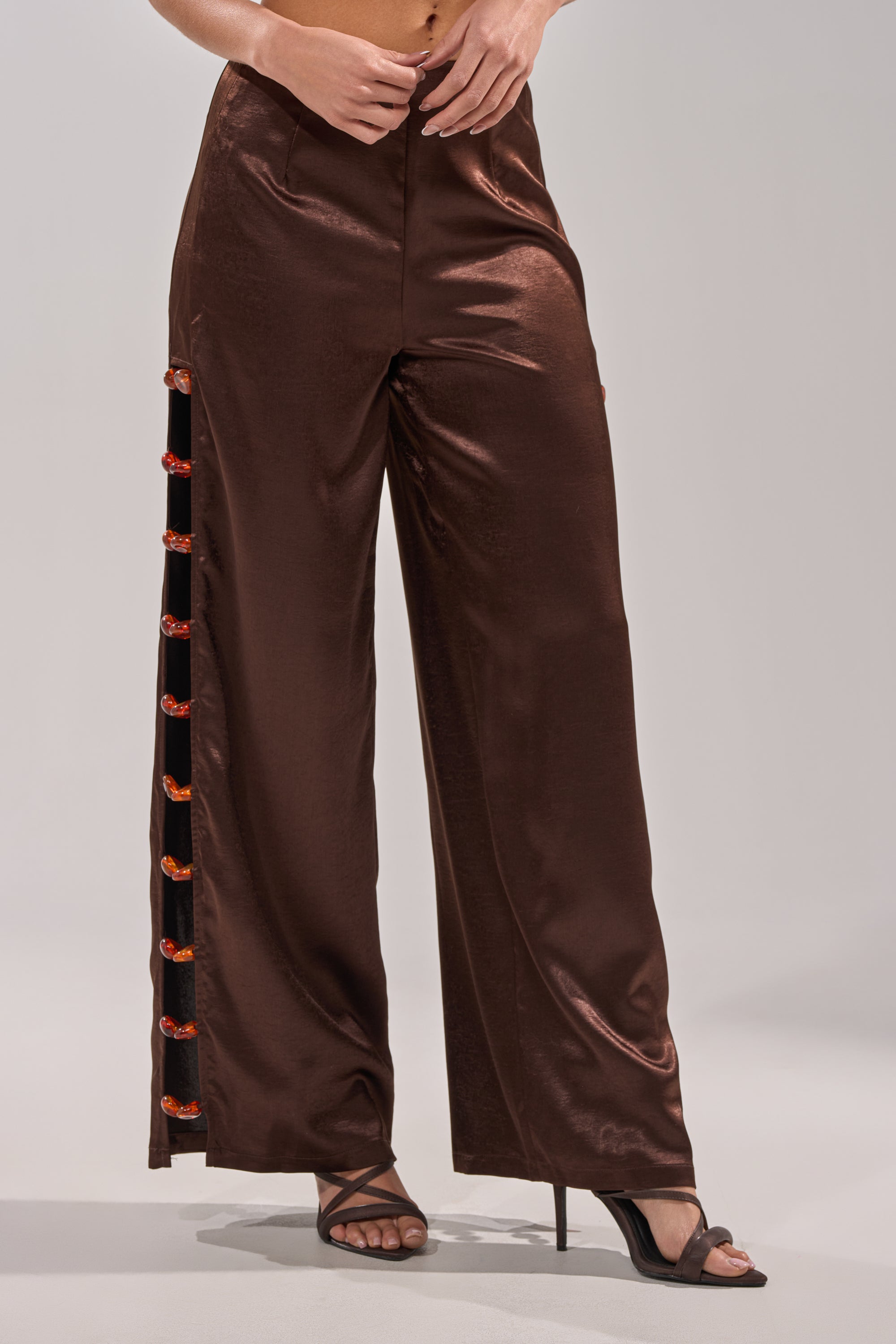 Someone is wearing the RESORT VIBES TROUSER—high-waisted, shiny brown satin wide-leg pants featuring red and orange accents on the left leg seam—paired with black open-toe high heel sandals. 2