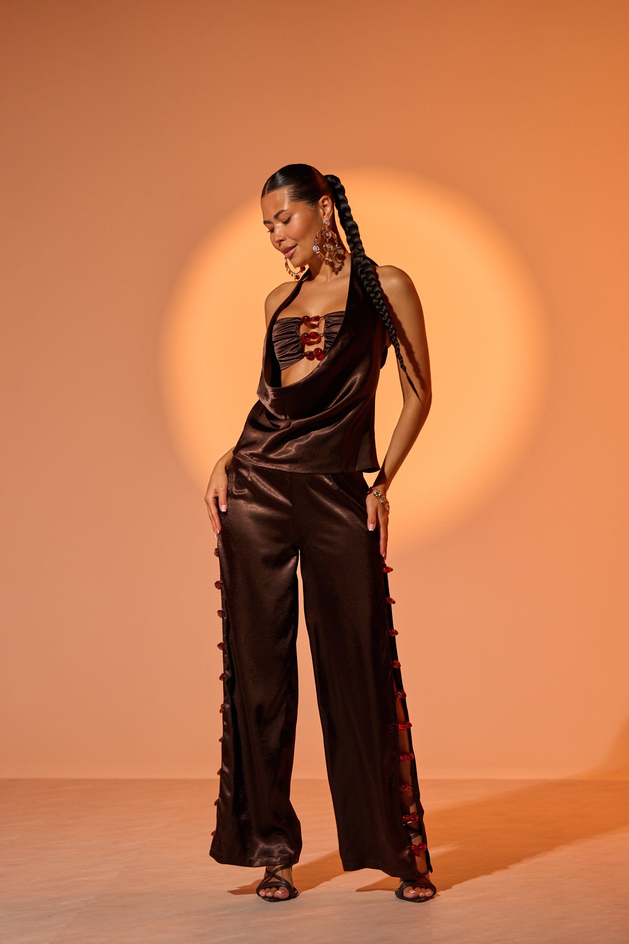 A woman stands confidently in a warm, orange spotlight, wearing the RESORT VIBES TROUSER—sleek dark brown satin wide-leg pants with side cutouts—paired with a matching halter top, statement earrings, and braided hair. 0