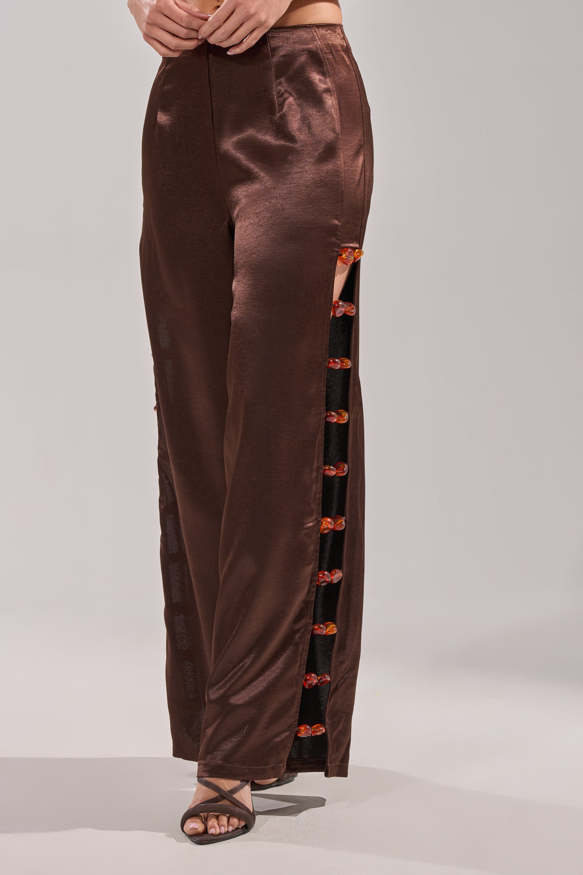 A person wearing the RESORT VIBES TROUSER—high-rise brown satin wide-leg pants with a side slit and red-orange frog closures—paired with black strappy heeled sandals for easy resort style. 3
