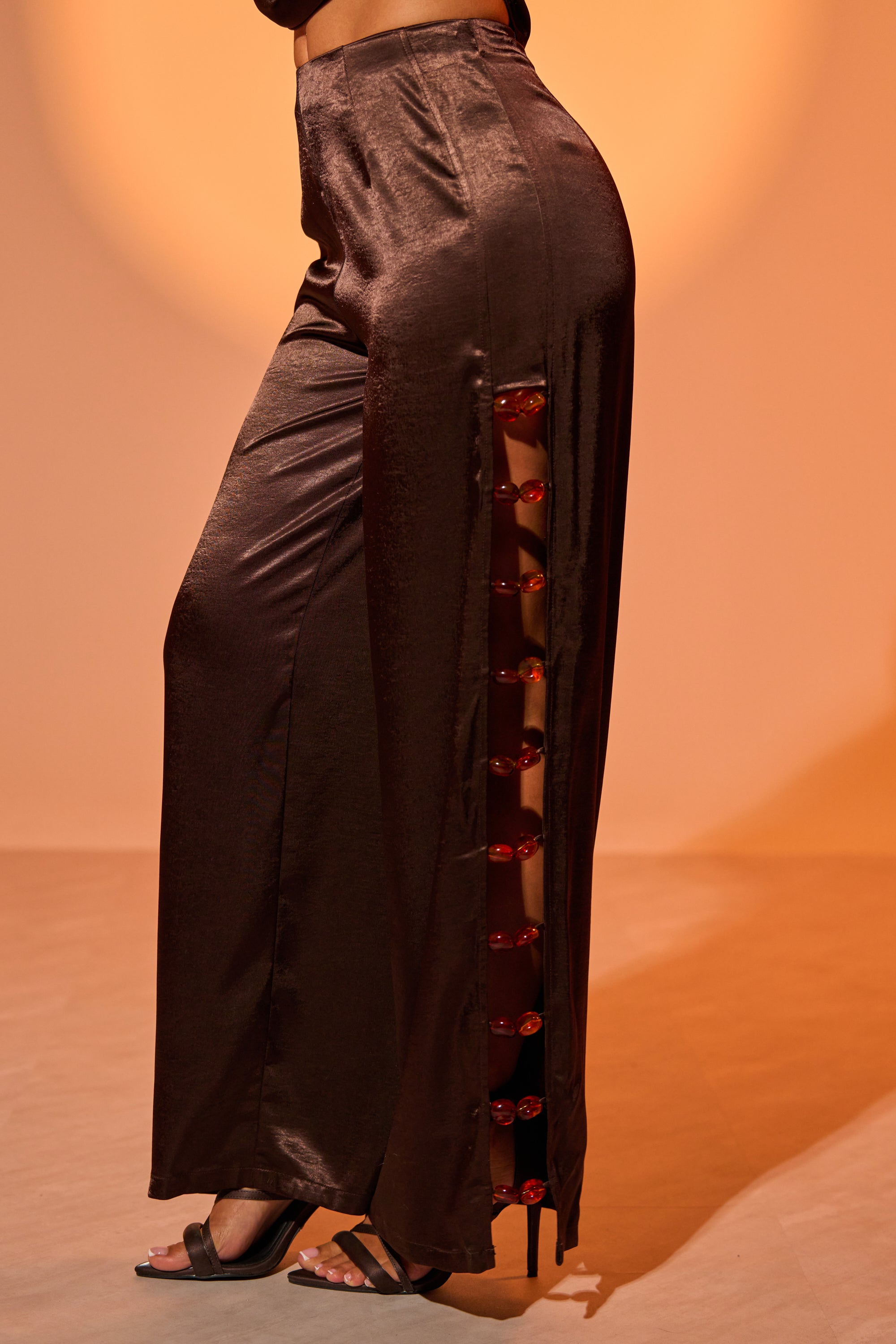 A person wears the shiny RESORT VIBES TROUSER featuring high-rise and side cutouts laced with red ribbon, paired with black heeled sandals. The softly lit, warm-toned background enhances the stylish look. 1