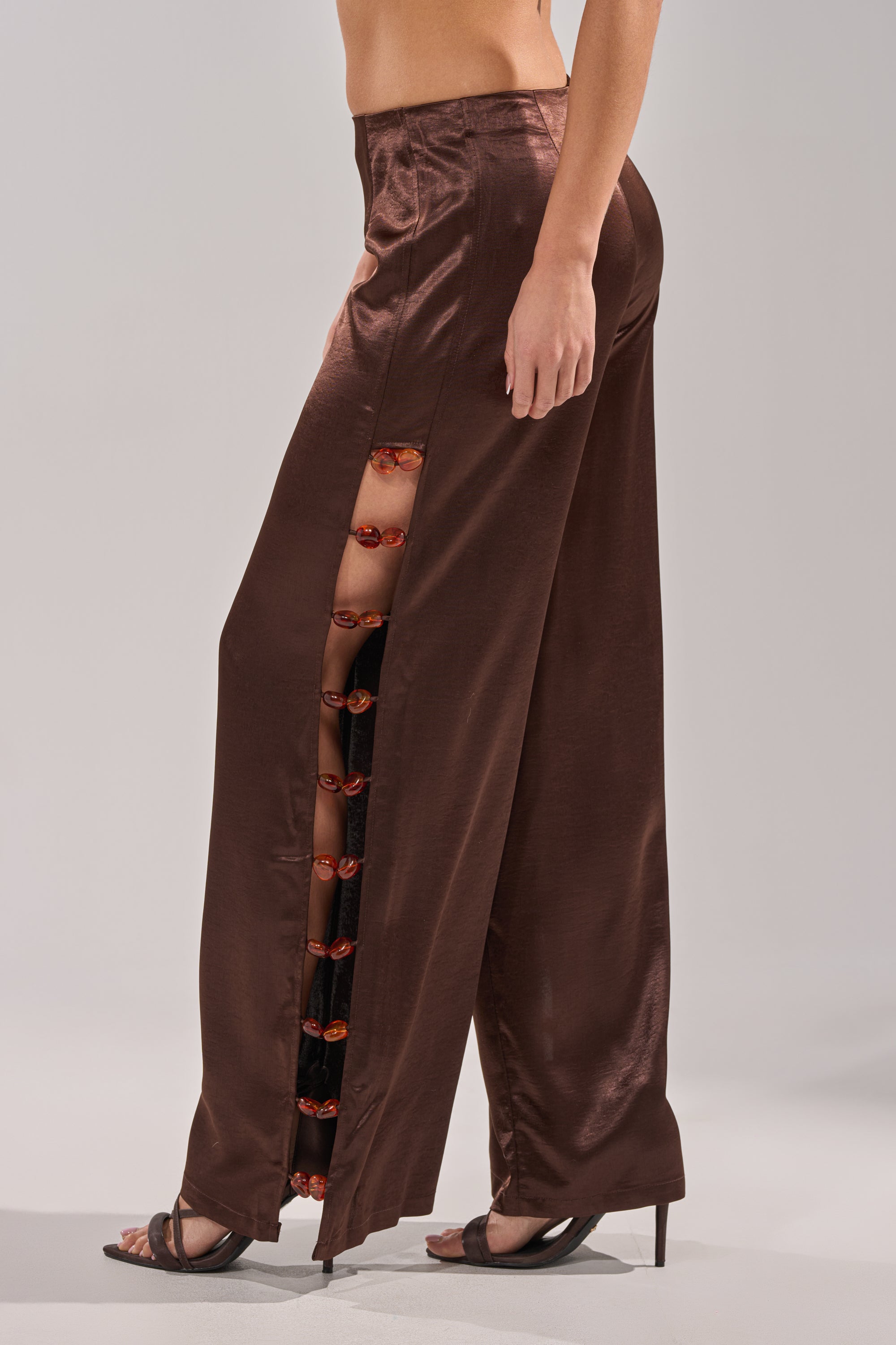 A person wears the RESORT VIBES TROUSER—high-rise brown satin wide-leg pants with open side seams and red buttons—paired with high-heeled sandals against a plain, light-colored background. 4