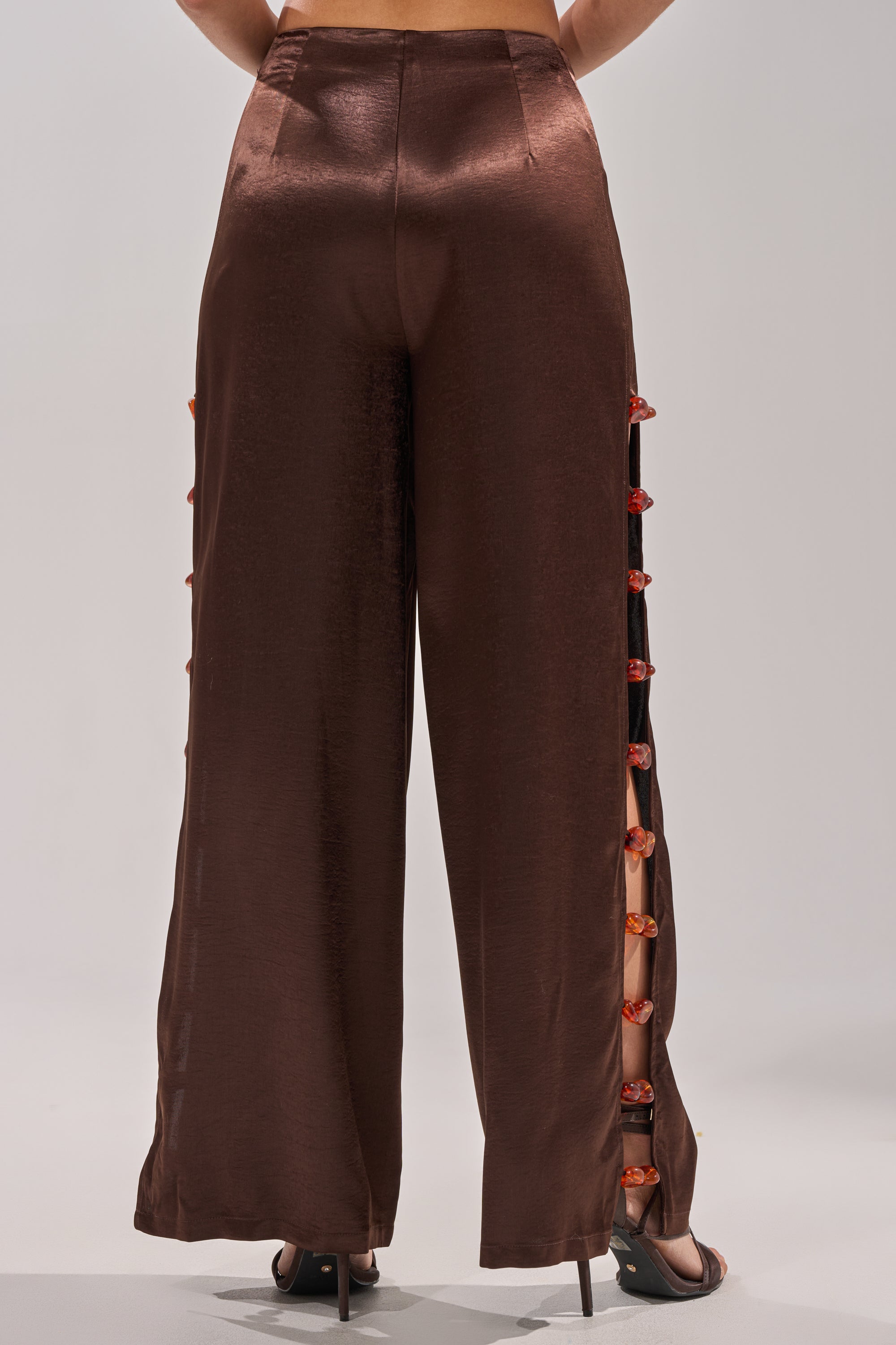A person is shown from the back against a plain background, wearing the shiny brown RESORT VIBES TROUSER, featuring red decorative buttons down the side seams and paired with open-toe heels. 5