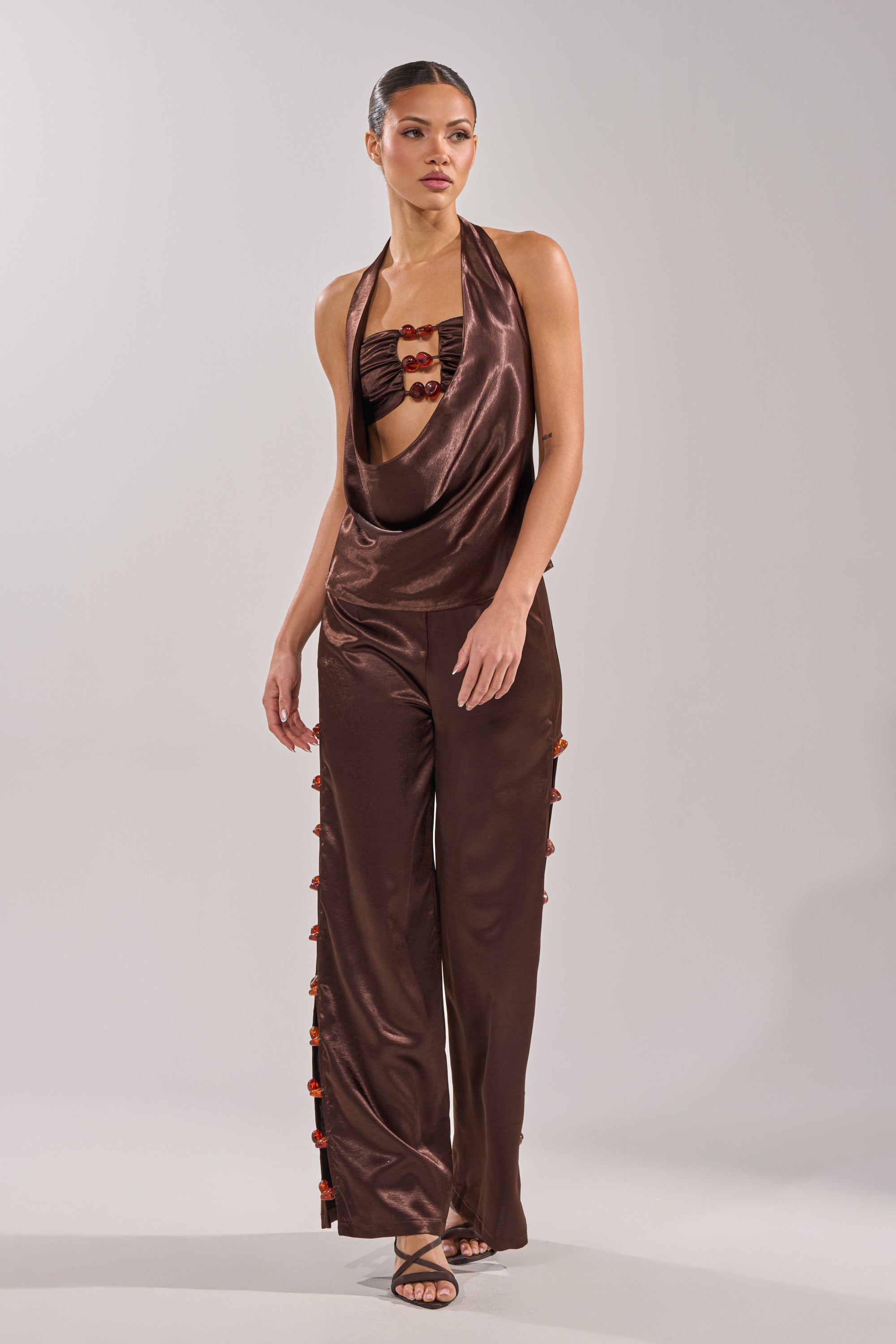 A woman models the RESORT VIBES TROUSER, a dark brown satin wide-leg pant, paired with a matching shiny halter top featuring a draped neckline and red accents. She completes the look with open-toe heels against a plain light background. 6