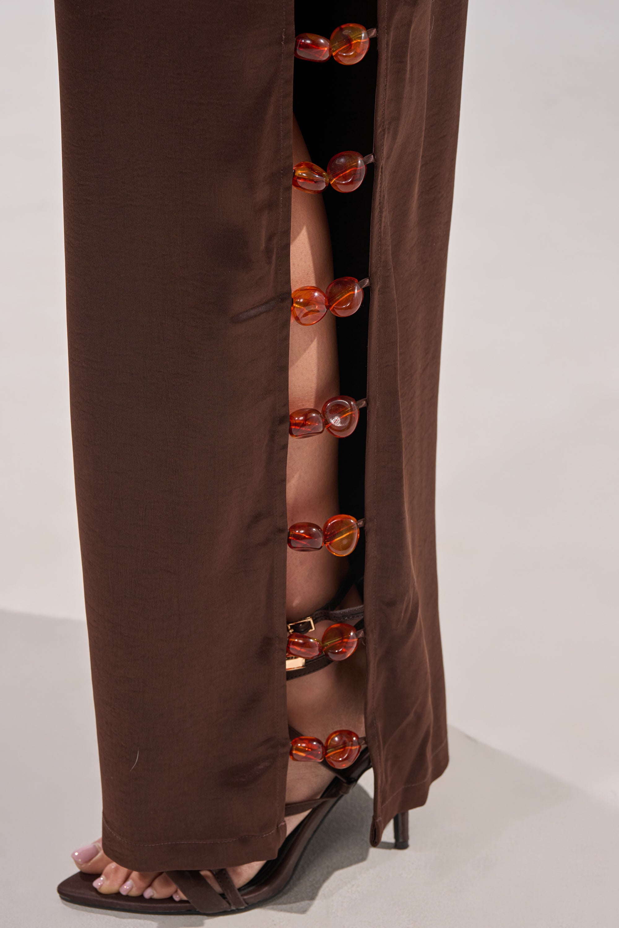A close-up of the RESORT VIBES TROUSER styled with a brown skirt featuring a high slit and large amber buttons, revealing part of a leg and sandal for effortless resort vibes. 9