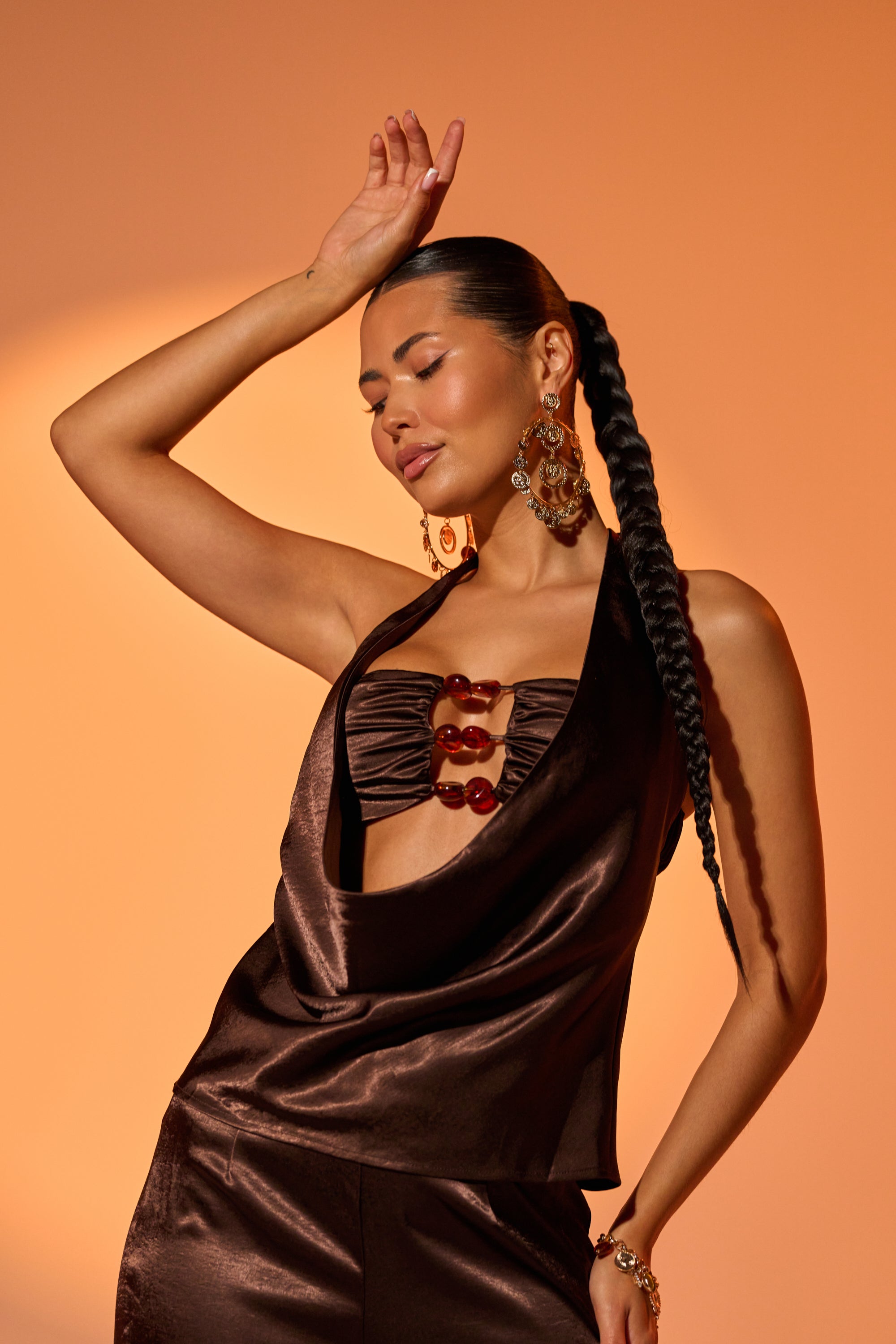 A woman with long, braided hair poses with one arm raised against an orange backdrop. She wears the RESORT VIBES BLOUSE—a halter satin top with red bead details—paired with matching pants, earrings, and a bracelet. 0