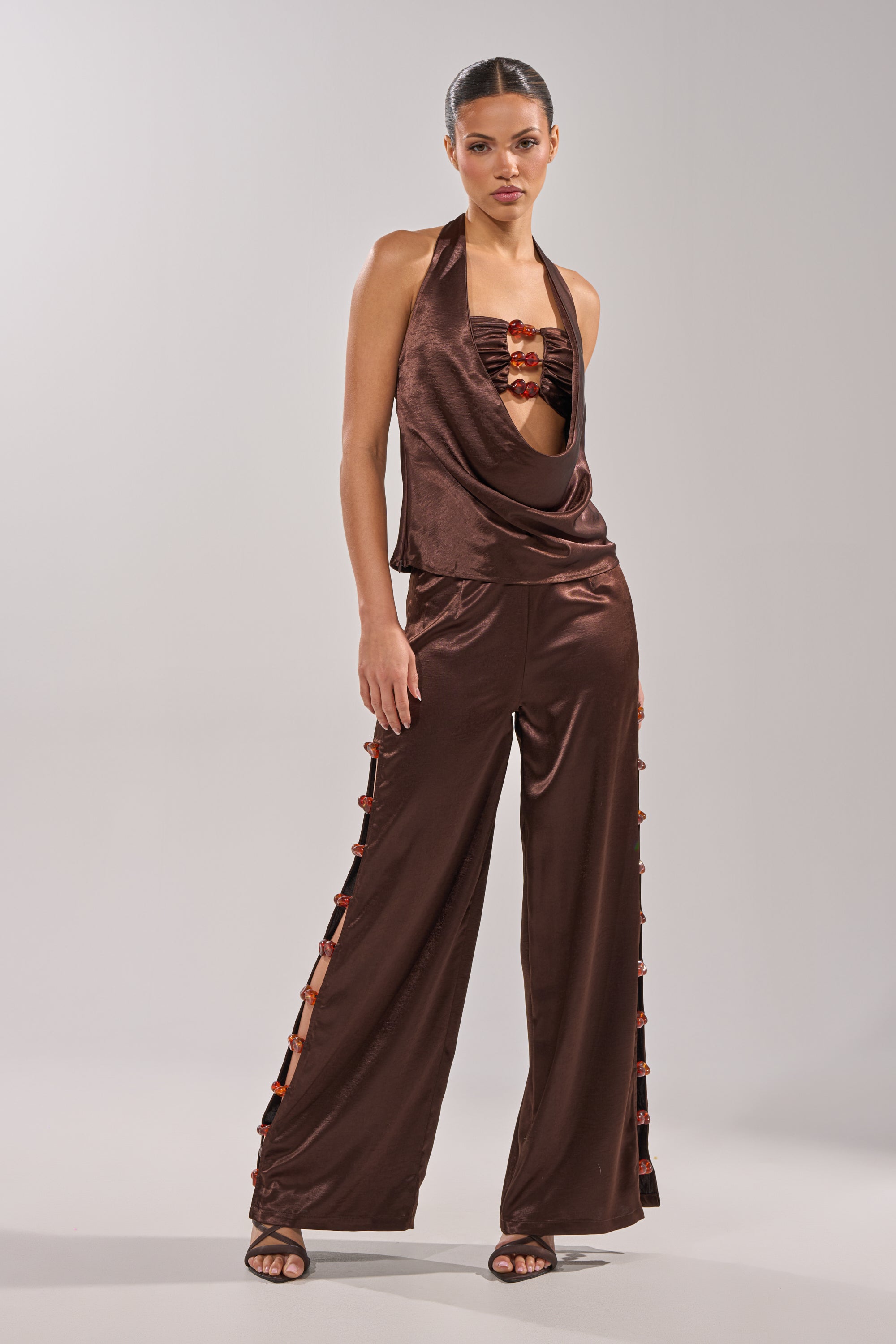 A woman models the RESORT VIBES BLOUSE, a satin cowl-front halter top in shiny brown, paired with matching wide-leg pants with side slits and red accents, styled elegantly with open-toe heels. 8