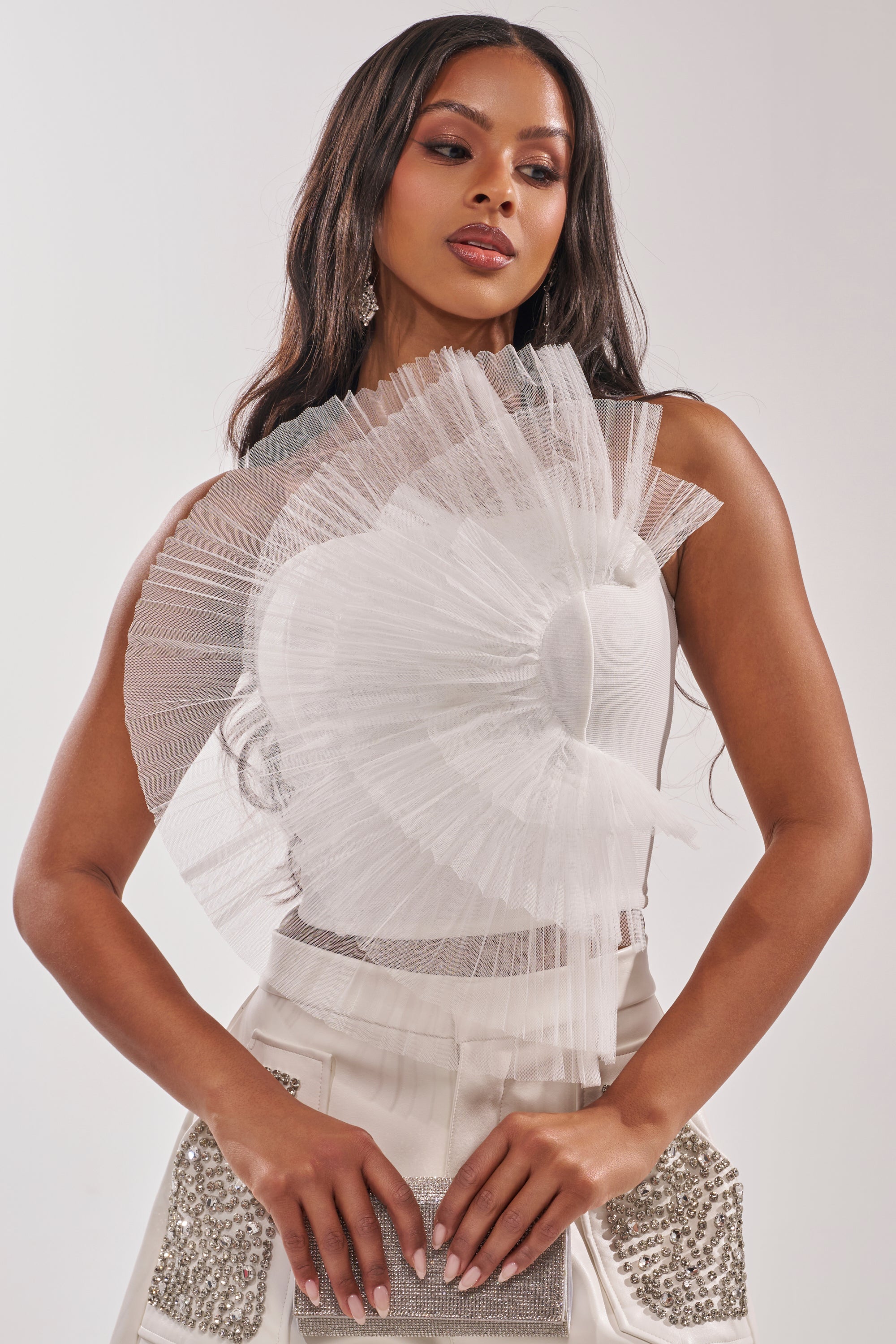 A woman with long, wavy dark hair wears the ALL DIFFERENT WAYS CORSET, a striking white top with a dramatic pleated tulle bodice. She holds a silver clutch and poses against a plain light background. 0