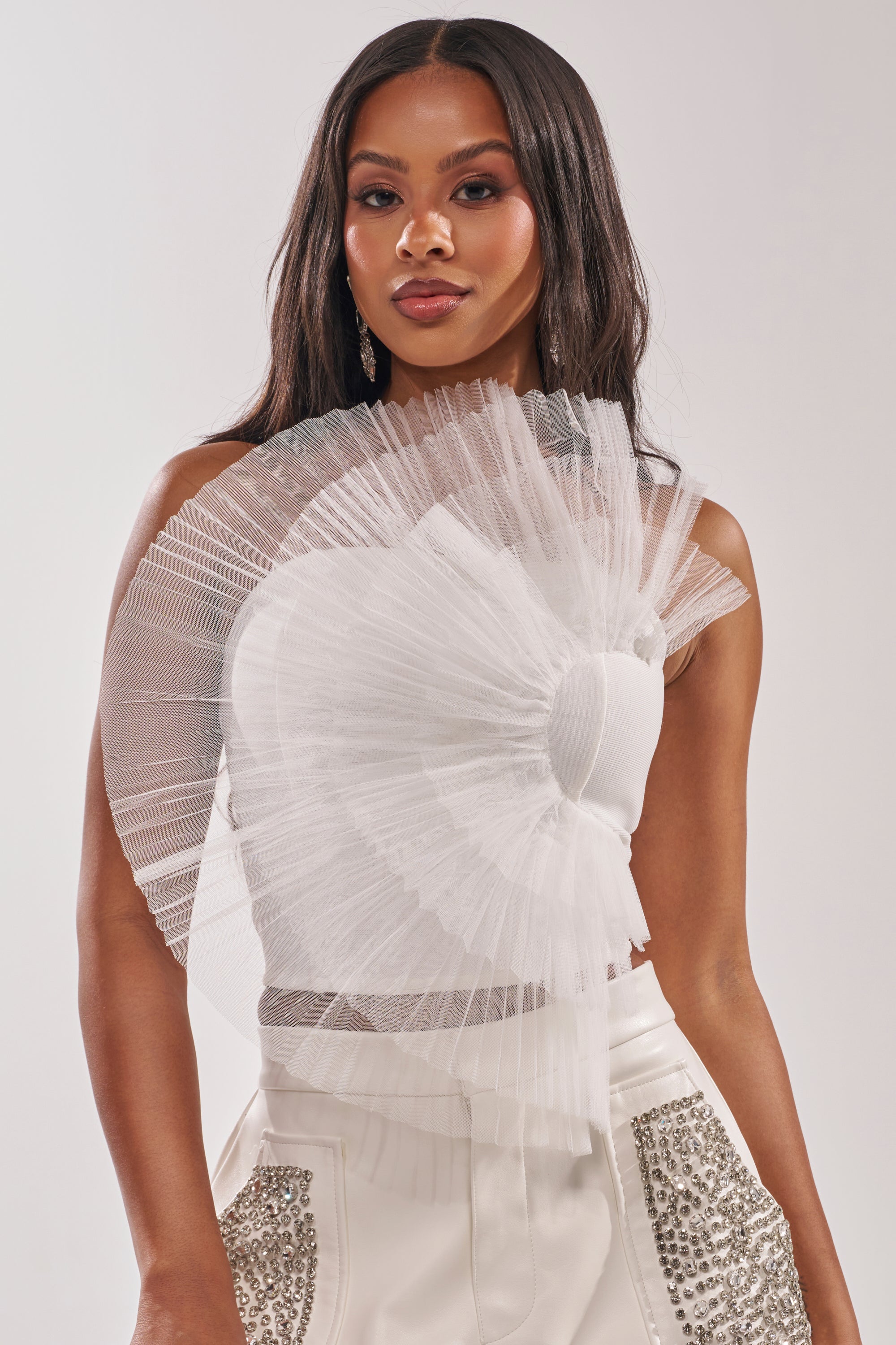 A woman wears the ALL DIFFERENT WAYS CORSET, an avant-garde white piece featuring dramatic pleated tulle ruffles over one shoulder, paired with white high-waisted silver-studded pants. She has long wavy hair and neutral makeup. 1