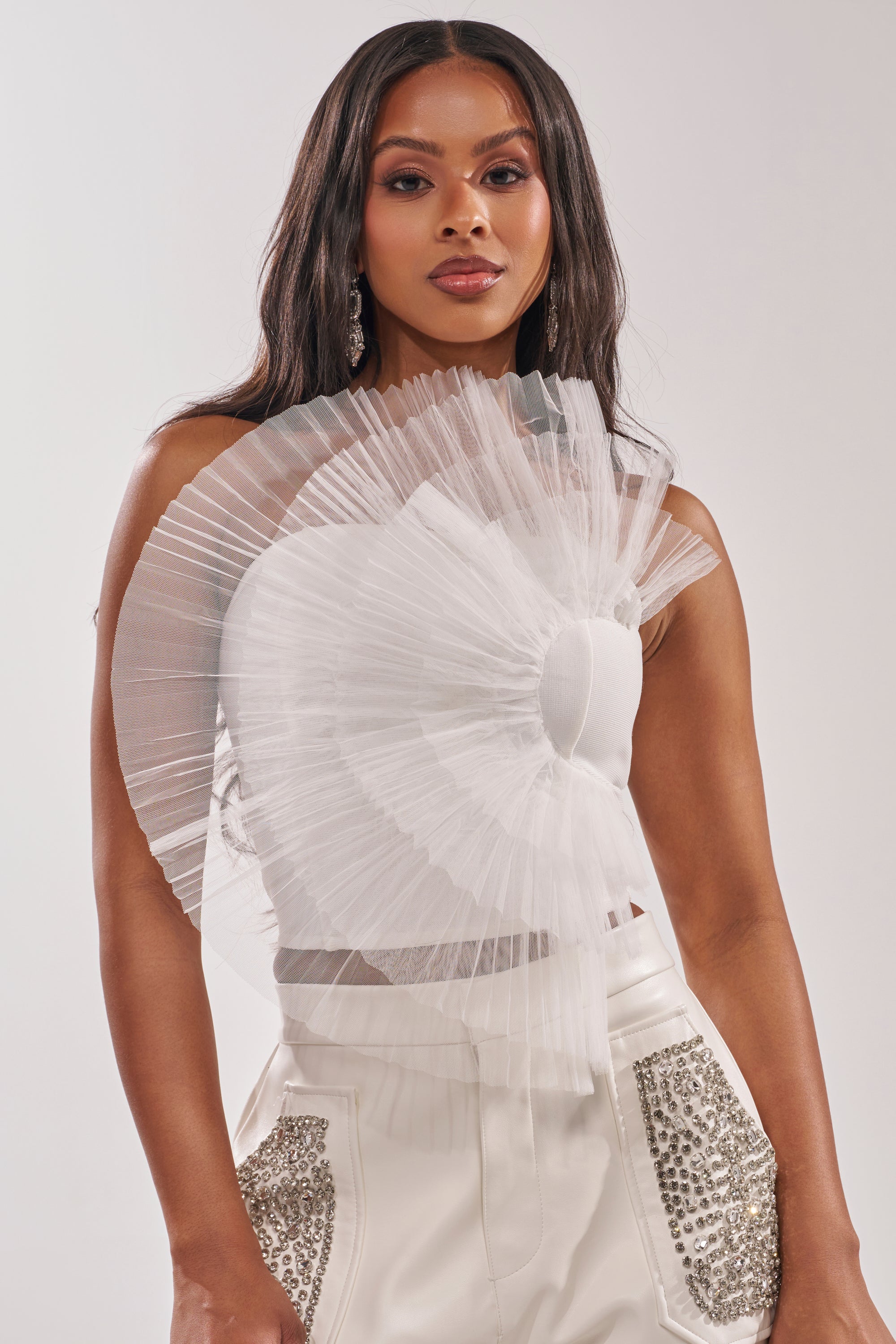 A woman wears the ALL DIFFERENT WAYS CORSET, a white top with dramatic pleated tulle on the shoulder and chest, paired with high-waisted white pants detailed with sparkling silver pocket embellishments. 2