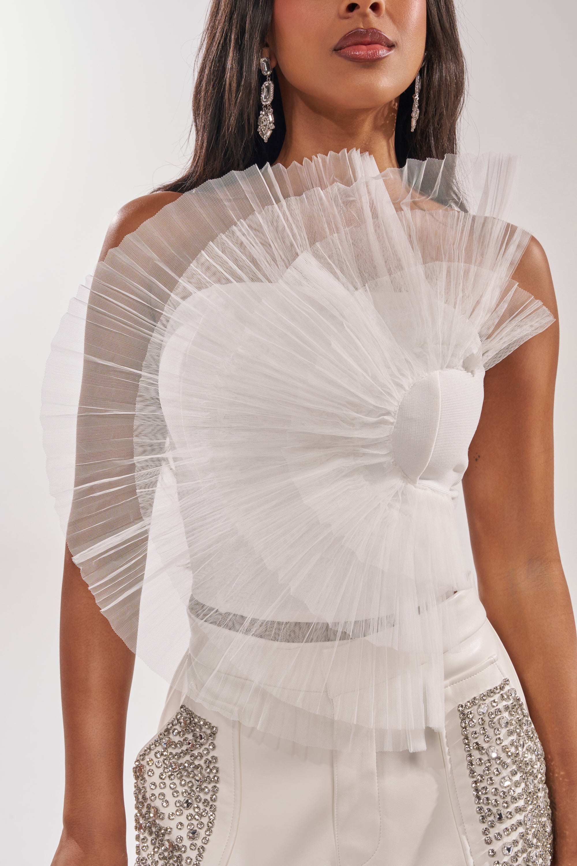 A woman wears the ALL DIFFERENT WAYS CORSET, a dramatic pleated tulle one-shoulder top, paired with high-waisted white pants featuring silver embellishments and finished with dangling earrings for an avant-garde look. 8