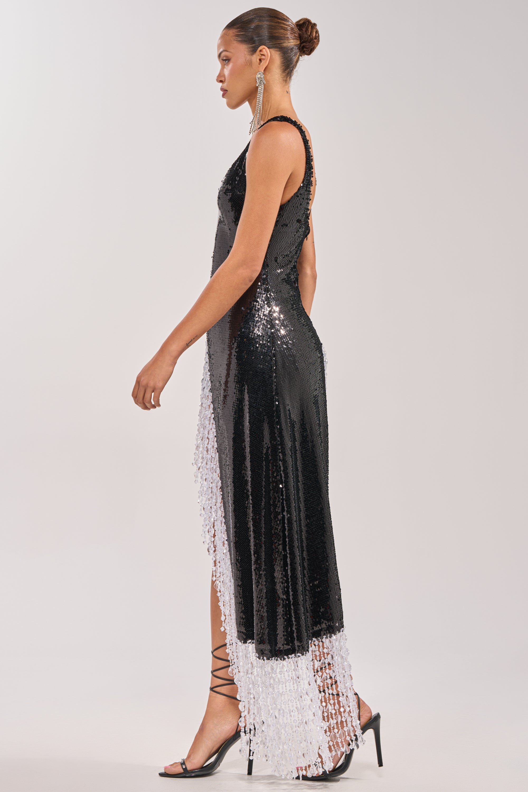 A woman in the NEW MONEY SEQUIN BLOUSE, a sleeveless black sequin top with an asymmetrical hem and white fringe beaded trim, paired with black strappy heels and statement earrings, stands sideways with her hair in a neat bun. 2