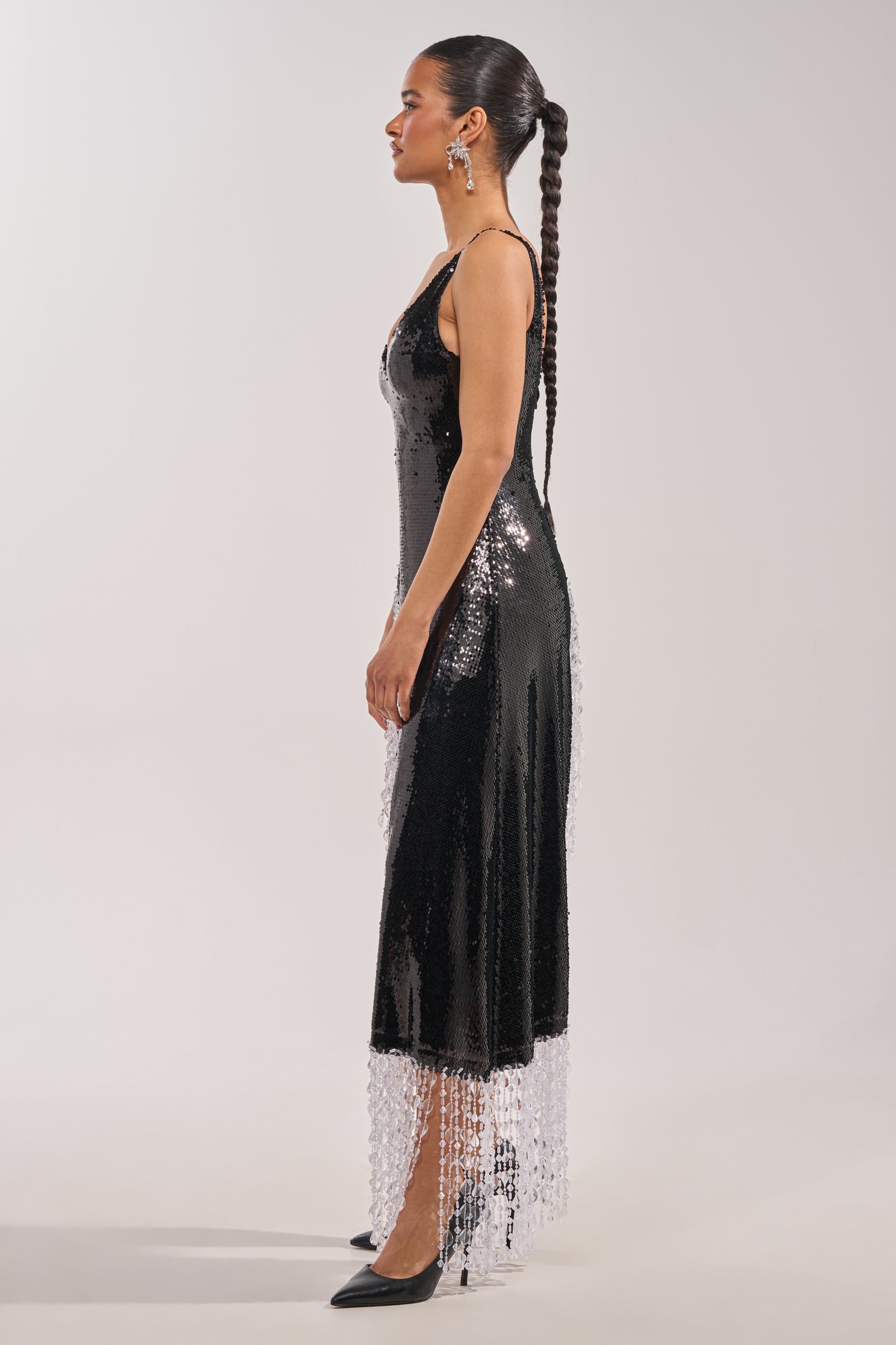 A woman with long braided hair stands in profile against a plain light background, wearing the NEW MONEY SEQUIN BLOUSE—sleeveless, black, covered in sequins with crystal-beaded fringe trim—and black high heels. 3