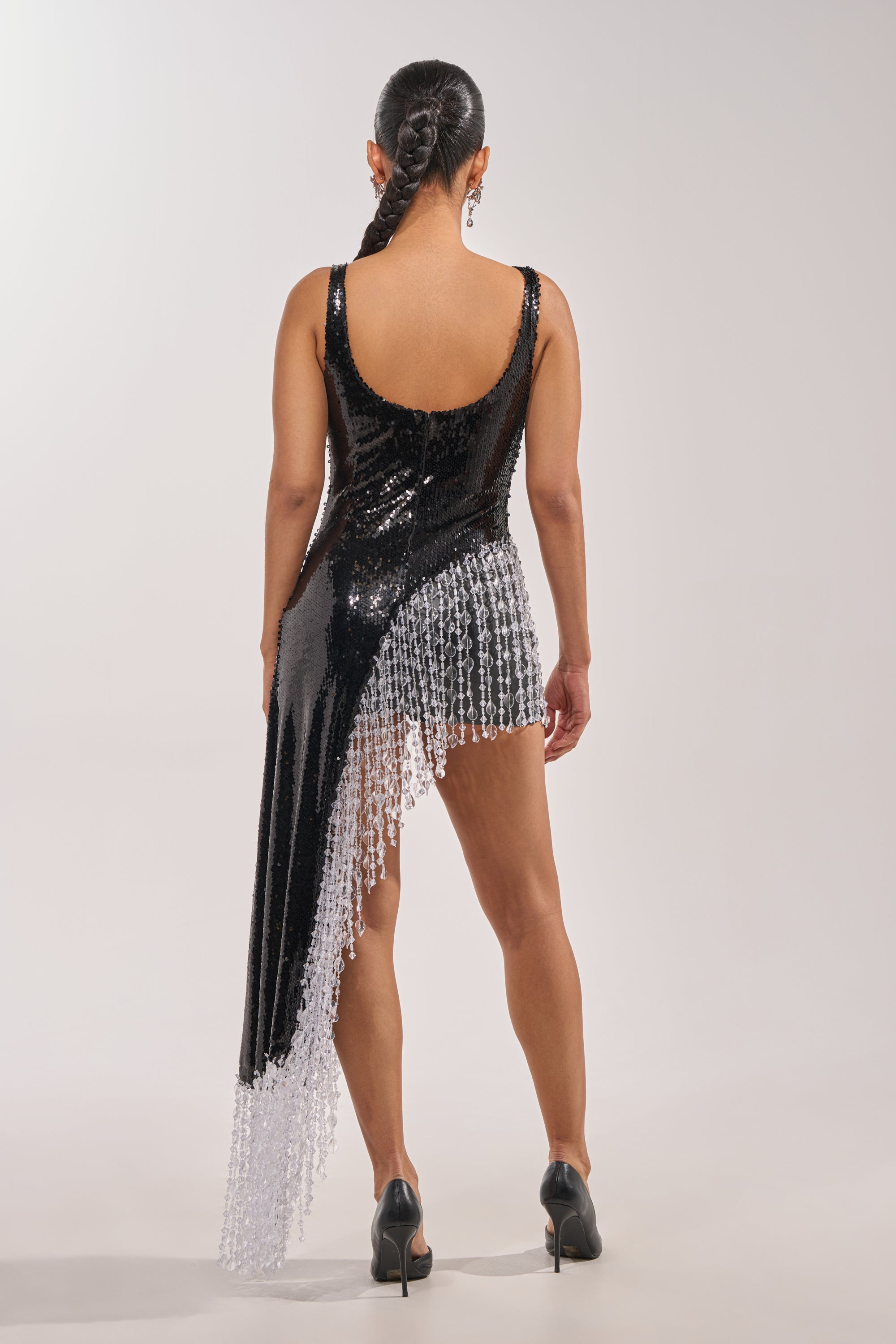 A woman stands with her back to the camera in the NEW MONEY SEQUIN BLOUSE, featuring black sequins and beaded fringe trim. She wears a sleek long braided ponytail and black heels against a plain light background. 5