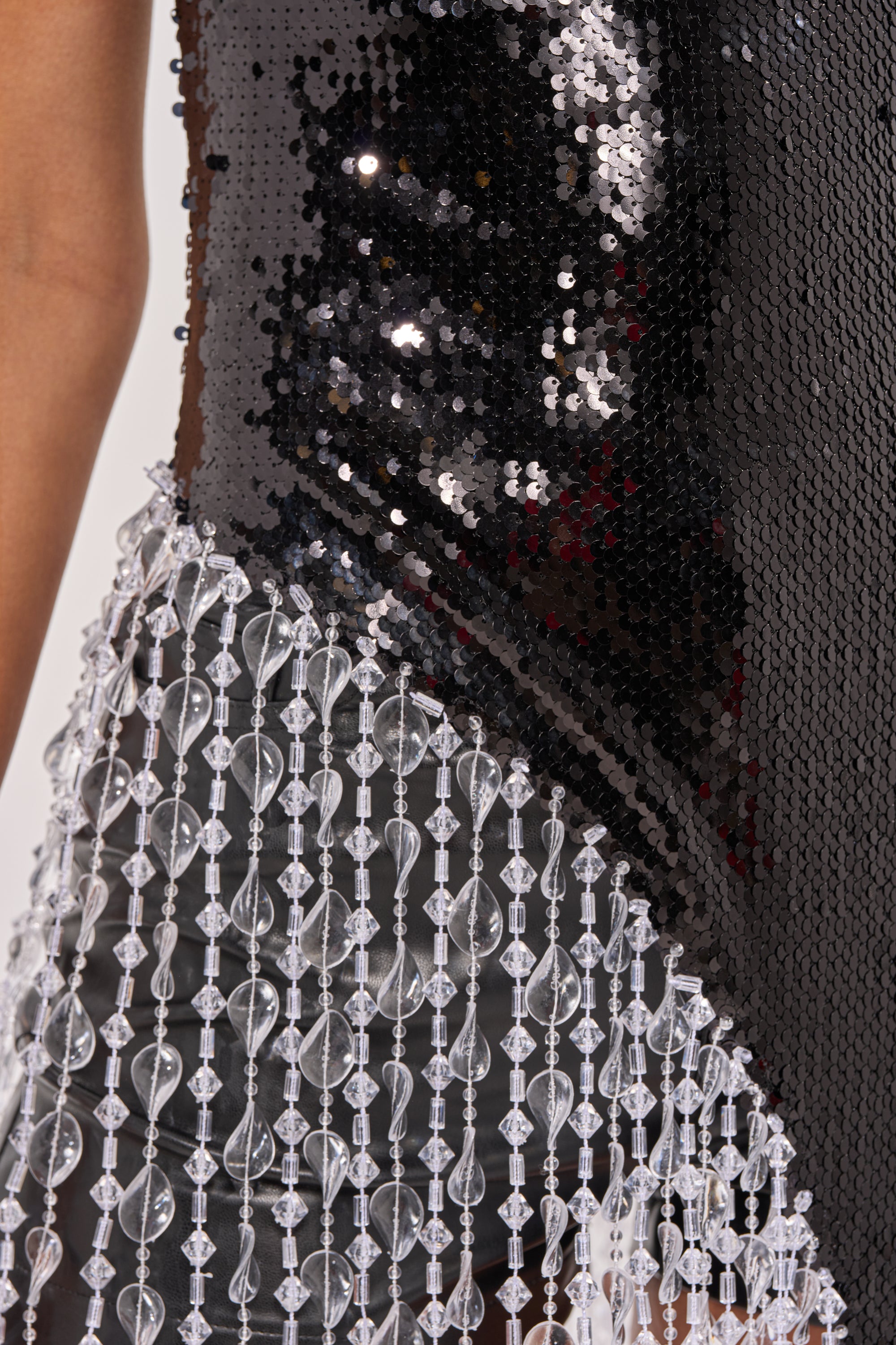 Close-up of the NEW MONEY SEQUIN BLOUSE, a black sequin top with clear bead and crystal fringe trim. The asymmetrical hem creates a sparkling, textured look. A person’s arm appears on the left side of the image. 5