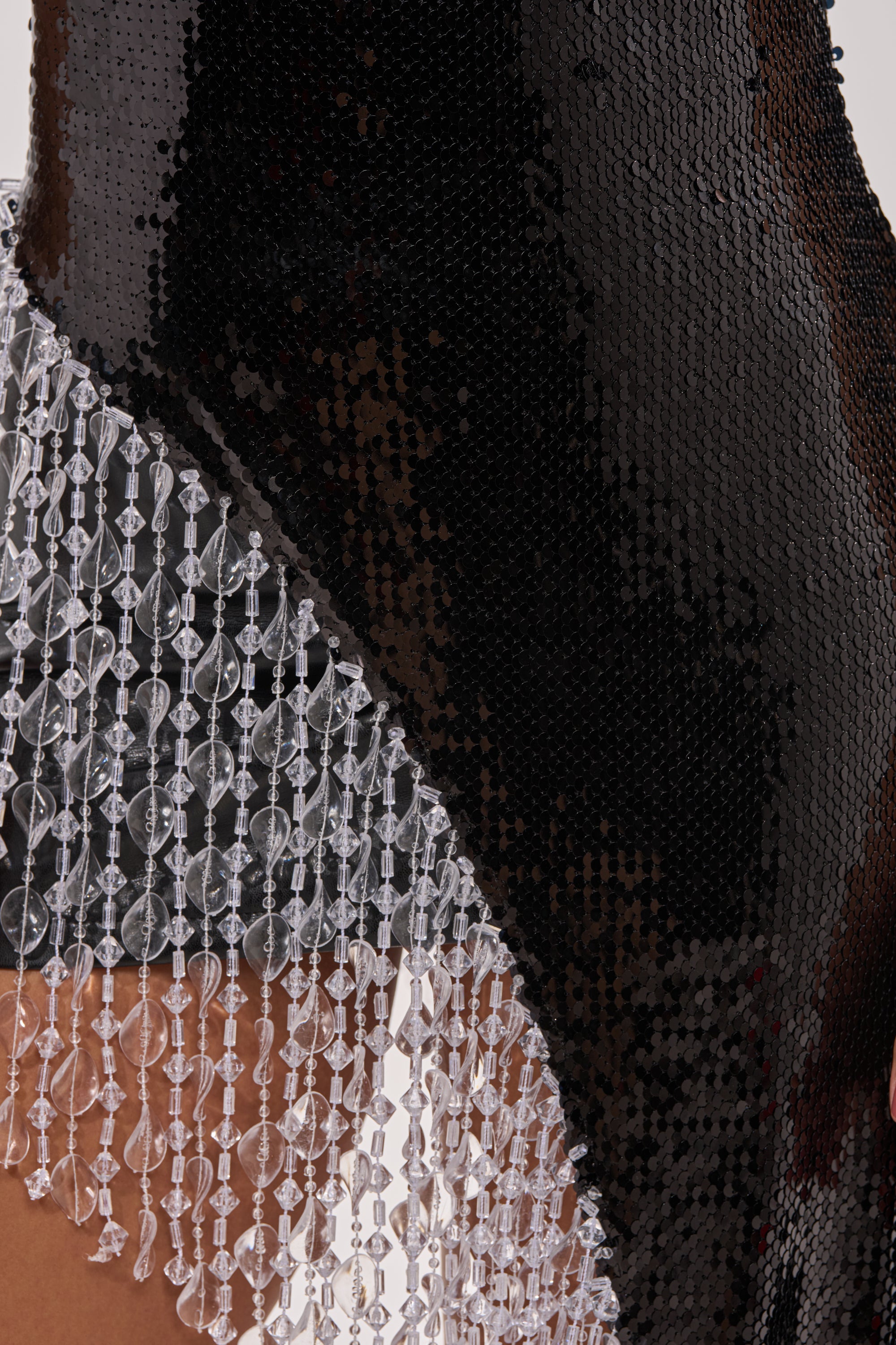 Close-up of the NEW MONEY SEQUIN BLOUSE, a black sheer sequin top with an asymmetrical hem and geometric-patterned clear bead fringe along the decorative edge. 6