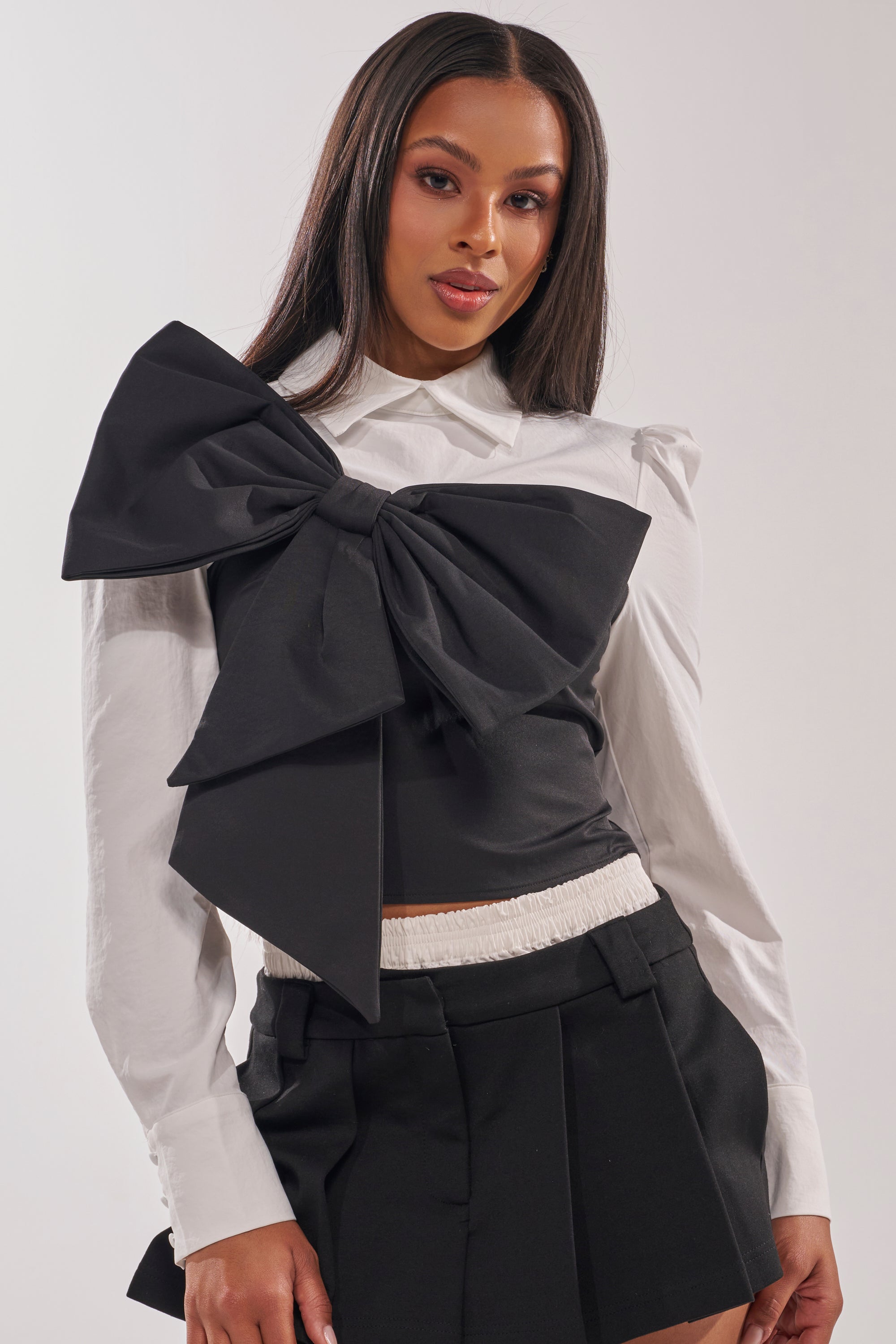 A woman wears the JACQUELINE POPLIN BLOUSE—a white long-sleeve top with a bold black bow—and black shorts featuring a white elastic waistband, posed against a plain light background. 0