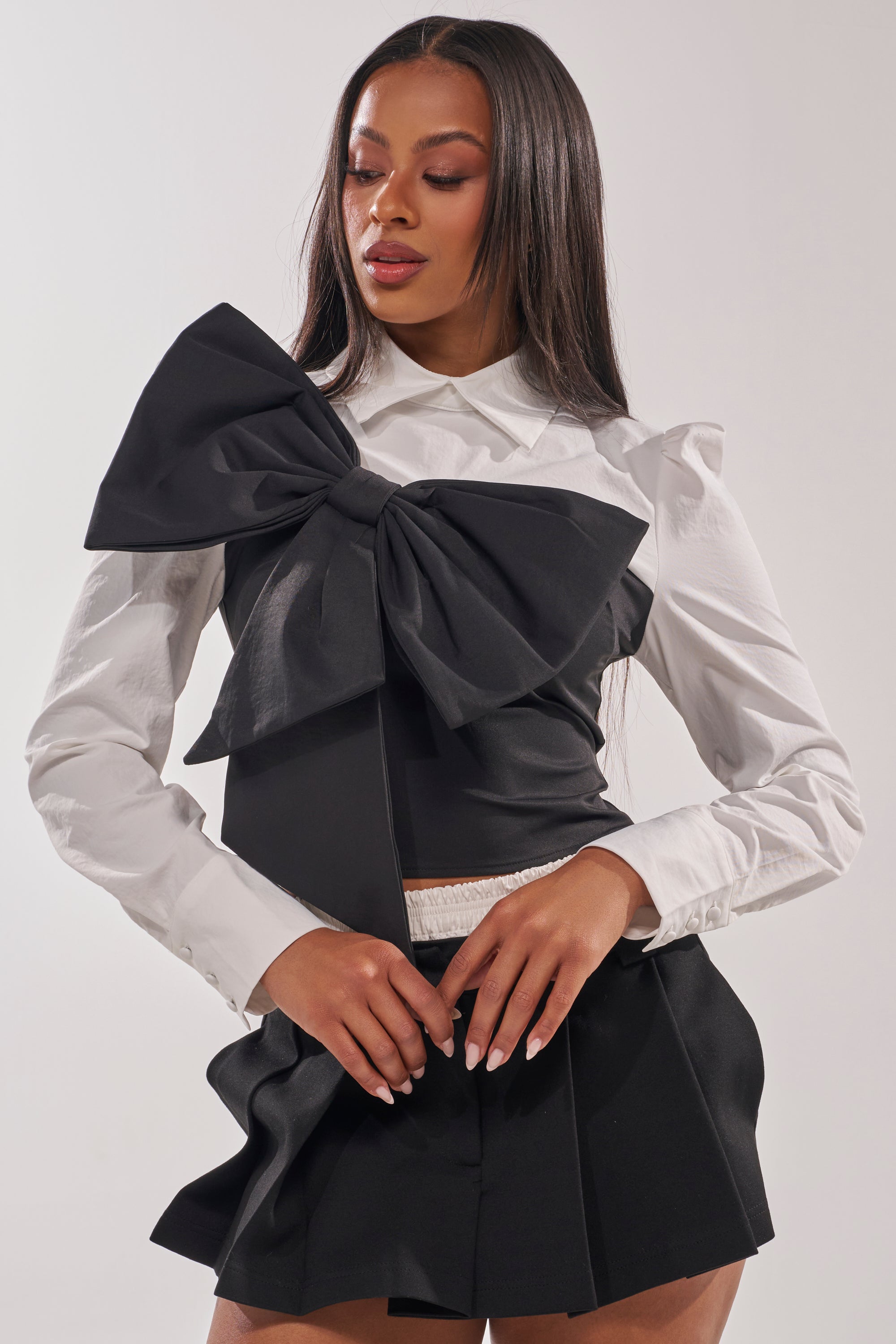 A woman stands against a plain light background, wearing the JACQUELINE POPLIN BLOUSE—a white long-sleeve top with a statement bow—paired with a black skirt. 1