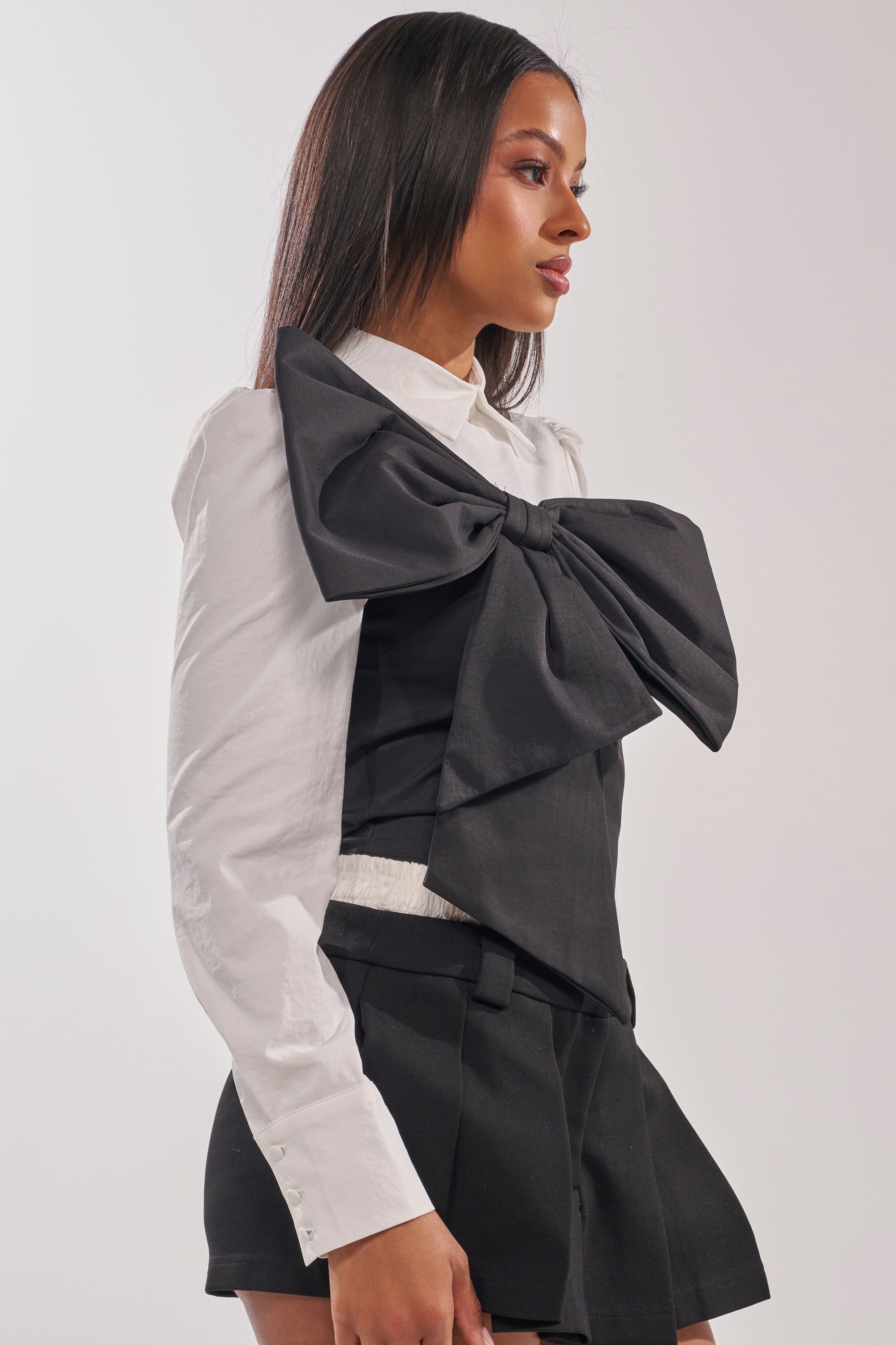 A woman with straight dark hair and a neutral expression models the JACQUELINE POPLIN BLOUSE, featuring a large black statement bow, paired with black shorts against a plain light background. 3