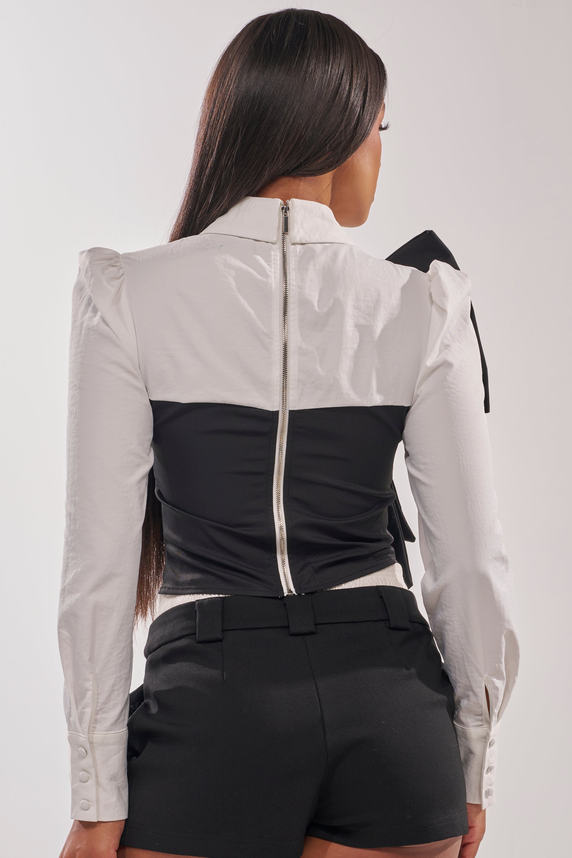 A woman with long straight hair, seen from the back, wears the JACQUELINE POPLIN BLOUSE featuring a white statement bow on one shoulder, a black waist section, visible silver zipper, and black fitted shorts. 4
