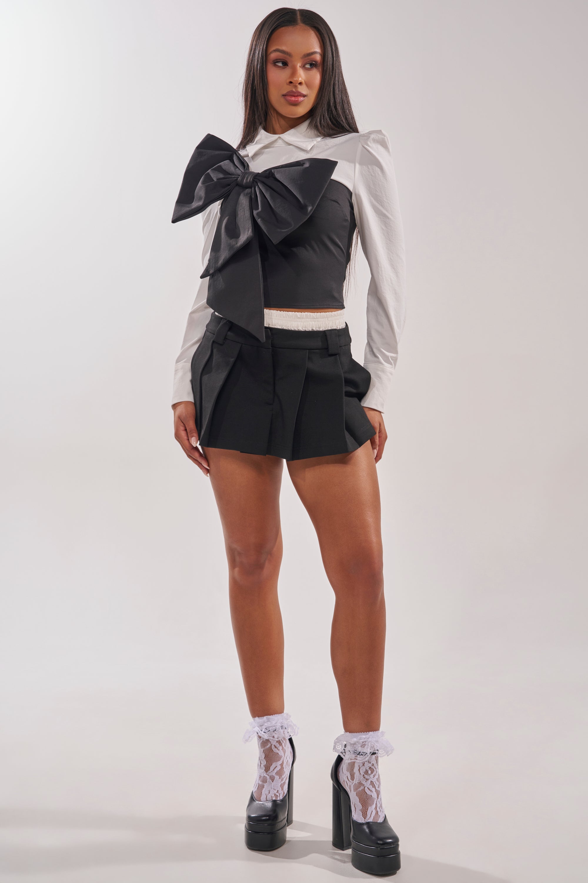 A woman faces forward against a plain background, wearing the JACQUELINE POPLIN BLOUSE with a white collar, black shorts, white lace socks, and black high-heeled platform shoes. 5
