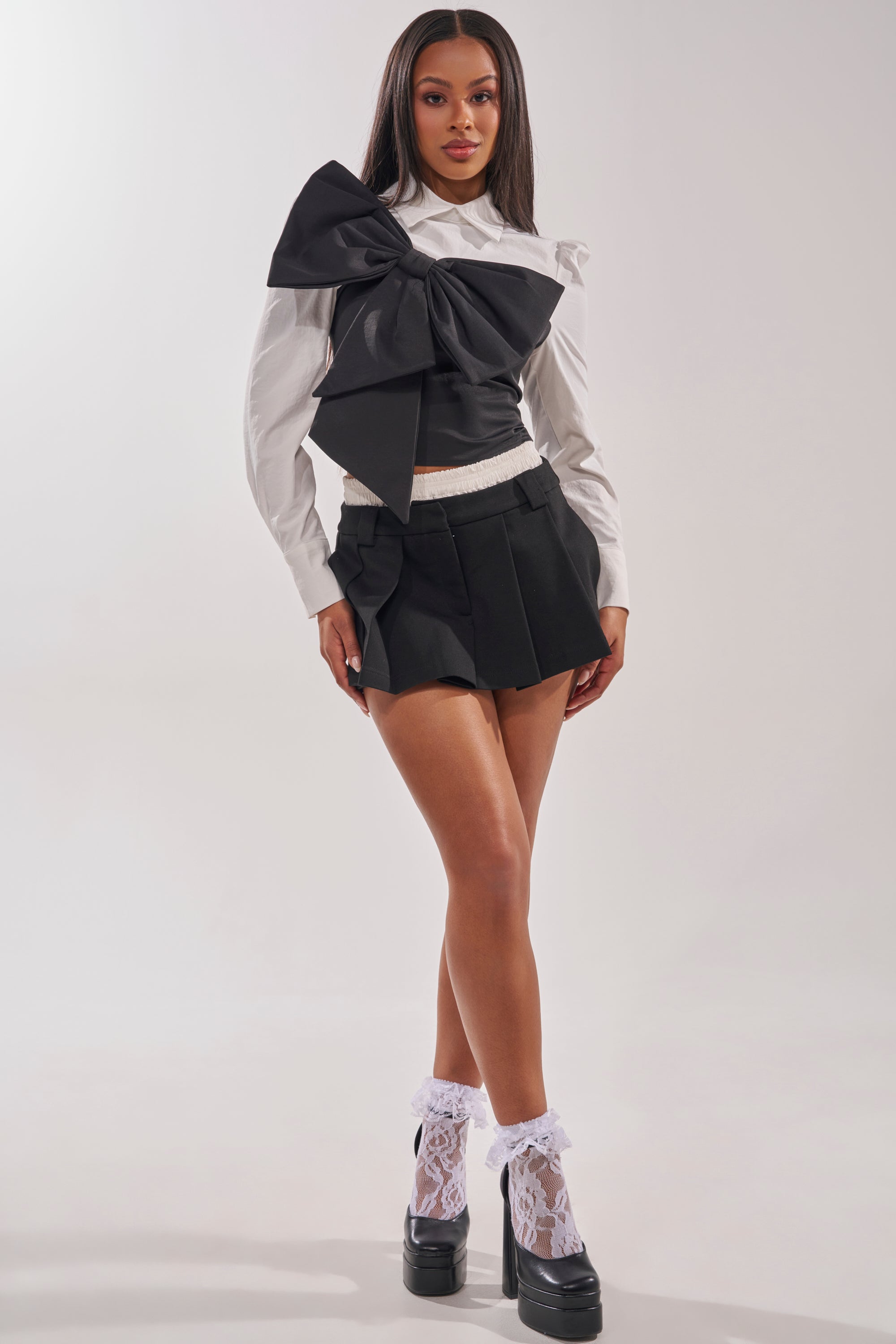 A woman stands confidently in the JACQUELINE POPLIN BLOUSE styled with a black mini skirt, white lace socks, and black platform shoes against a plain light background. 6