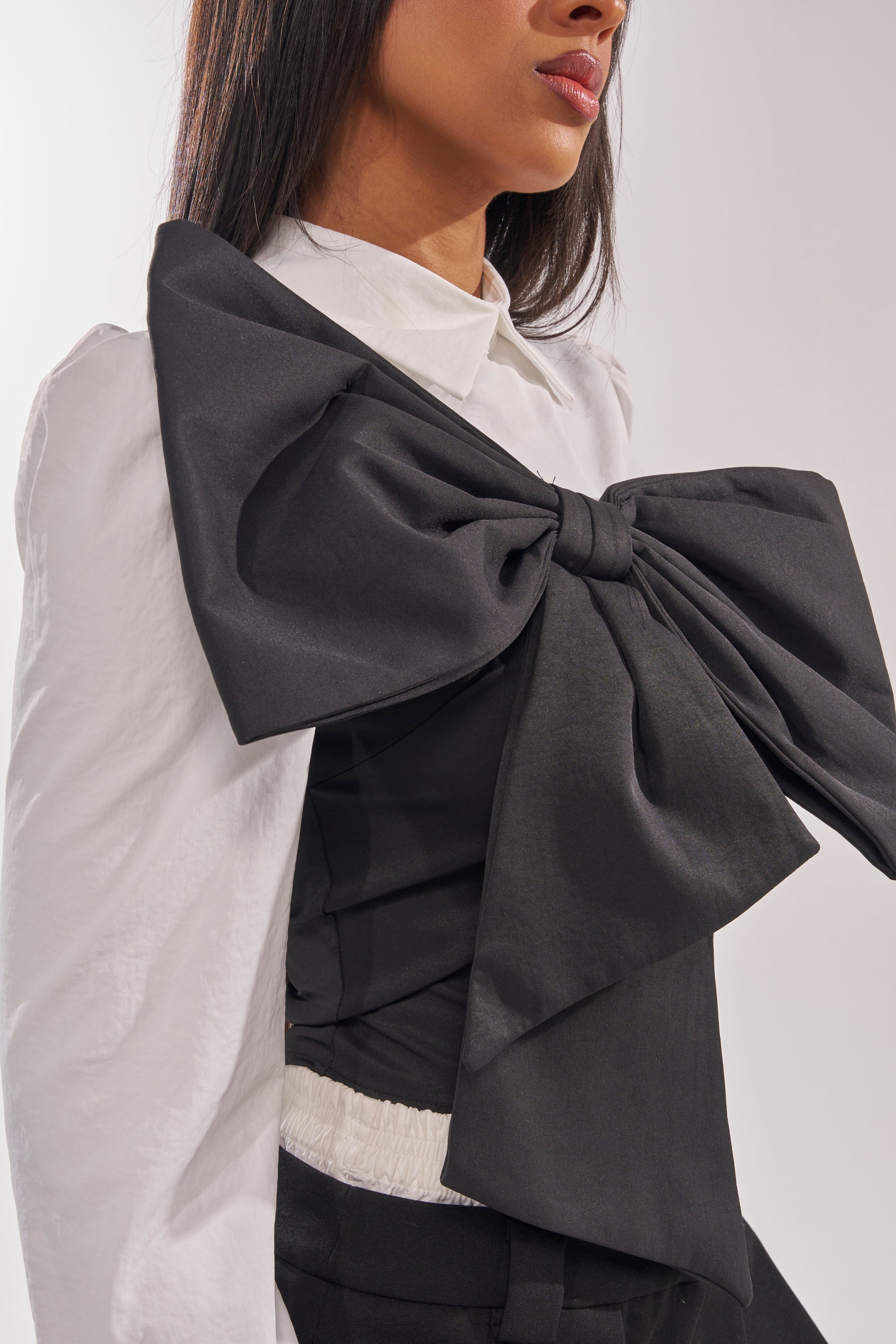 A woman wearing the JACQUELINE POPLIN BLOUSE, featuring a sculptural silhouette and a dramatic black bow over her shoulder and chest, stands against a plain background. 7
