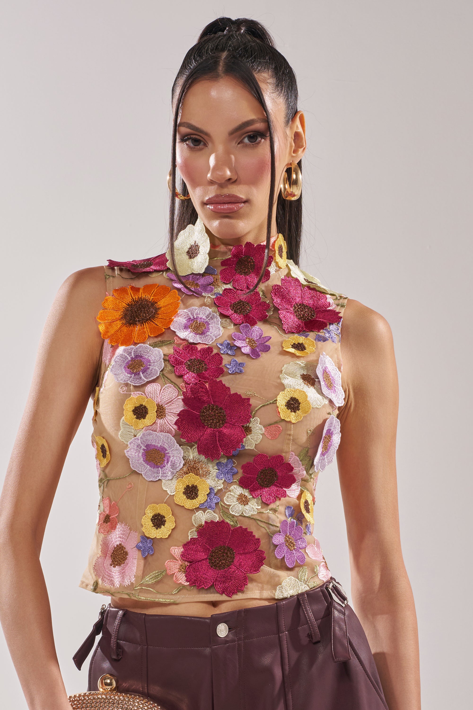 A woman poses against a plain light background wearing the WHERE THIS FLOWER BLOOMS MESH TOP—sleeveless, mock neck, sheer mesh with colorful 3D floral embroidery—paired with high-waisted maroon pants and gold hoop earrings. 0